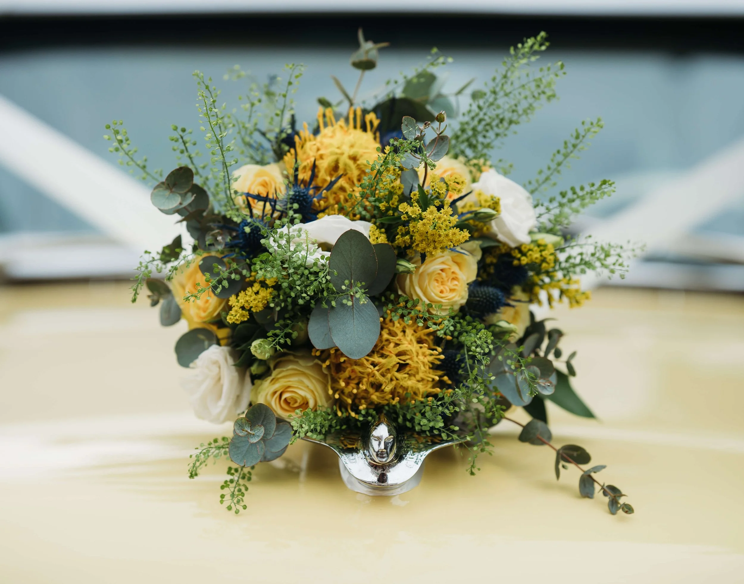 A floral arrangement with yellow roses, yellow chrysanthemums, white roses, yellow and green fillers, and eucalyptus leaves in a silver vase on a yellow table.