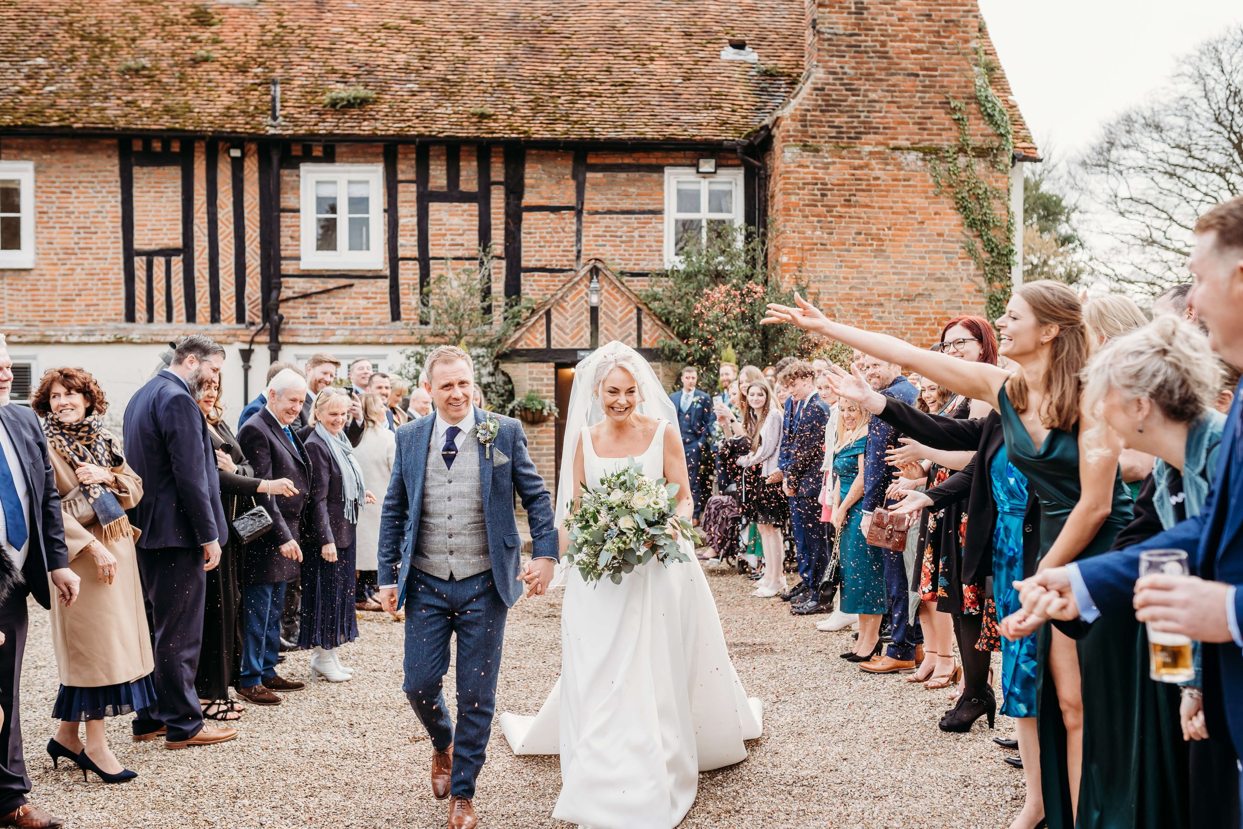 A bride and groom walking hand in hand at their wedding outdoors, surrounded by friends and family celebrating and throwing confetti.