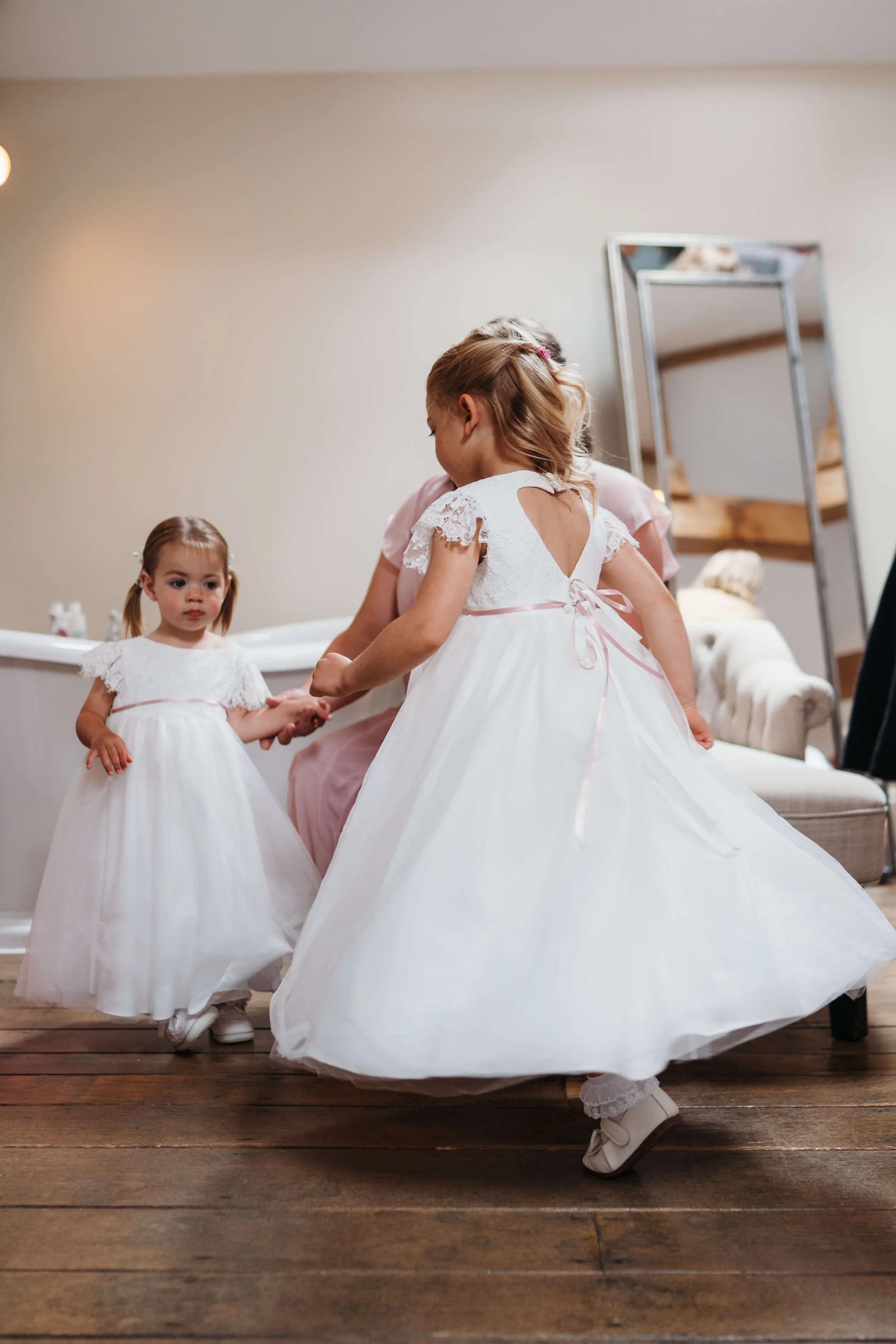 Three young girls in white dresses, possibly flower girl dresses, holding hands and dancing indoors on wooden floors.