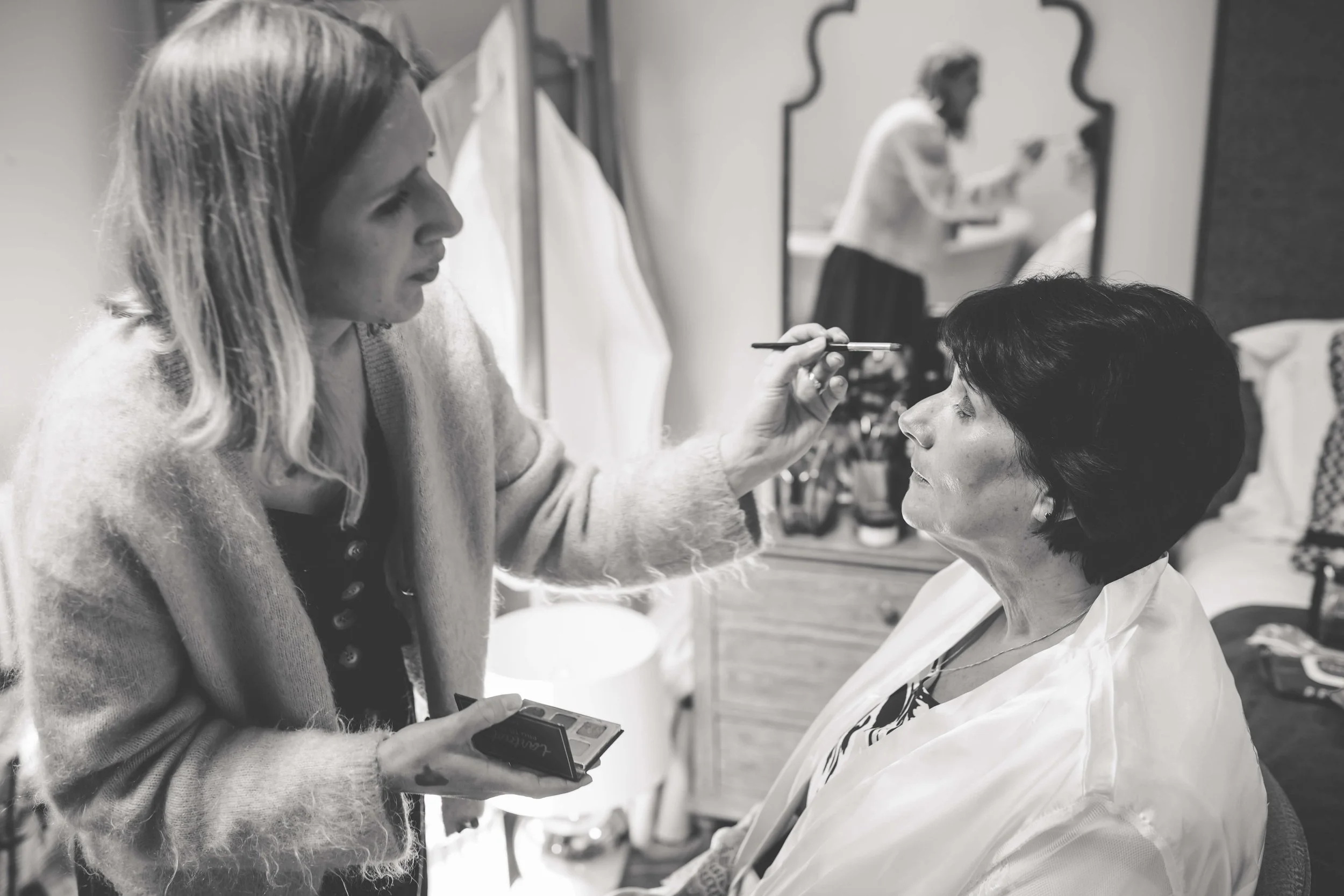 Makeup artist applying makeup to a woman sitting in a chair, with a mirror and makeup supplies in the background.
