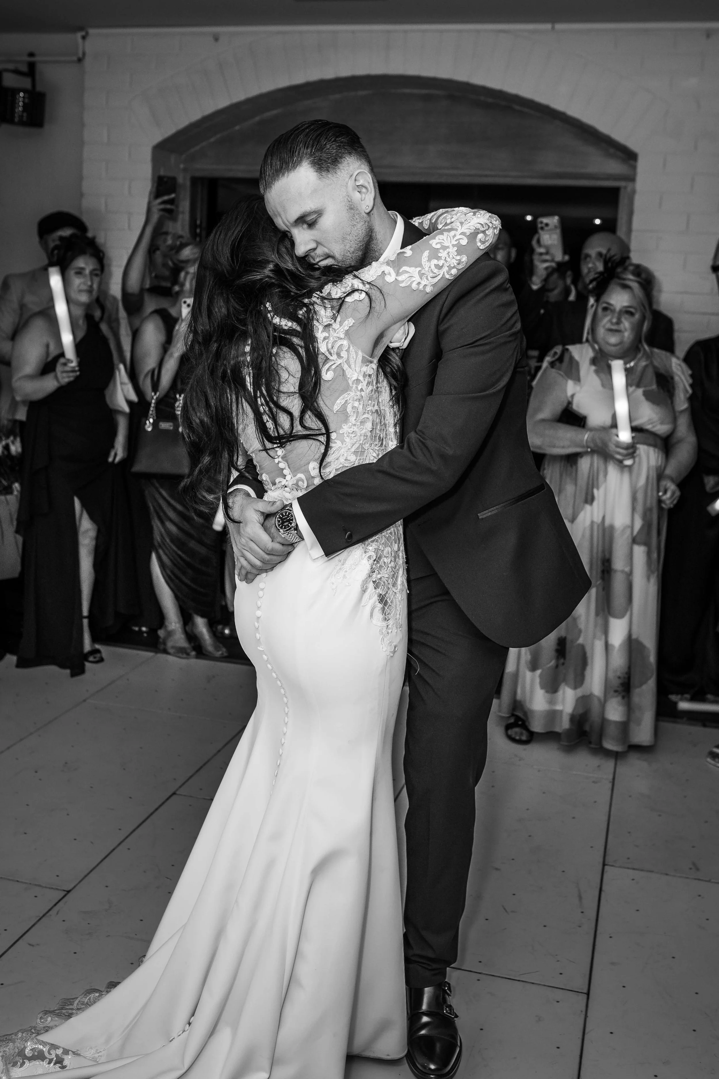 A black and white photo of a bride and groom sharing a dance at their wedding reception, surrounded by onlookers.