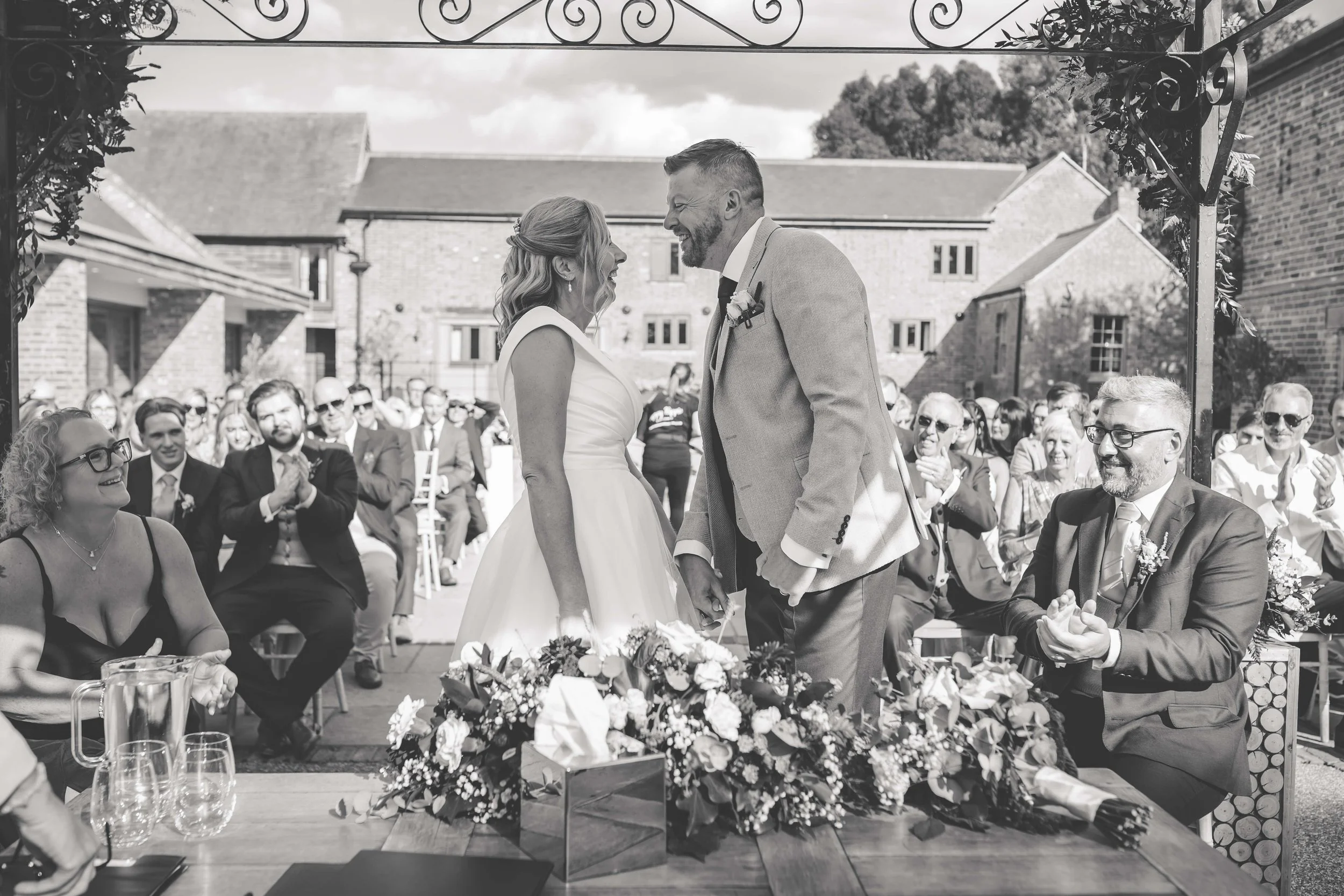 A wedding ceremony outdoors with a bride and groom smiling and holding hands, surrounded by seated guests clapping and smiling, with a flower arrangement on the table in front of them.