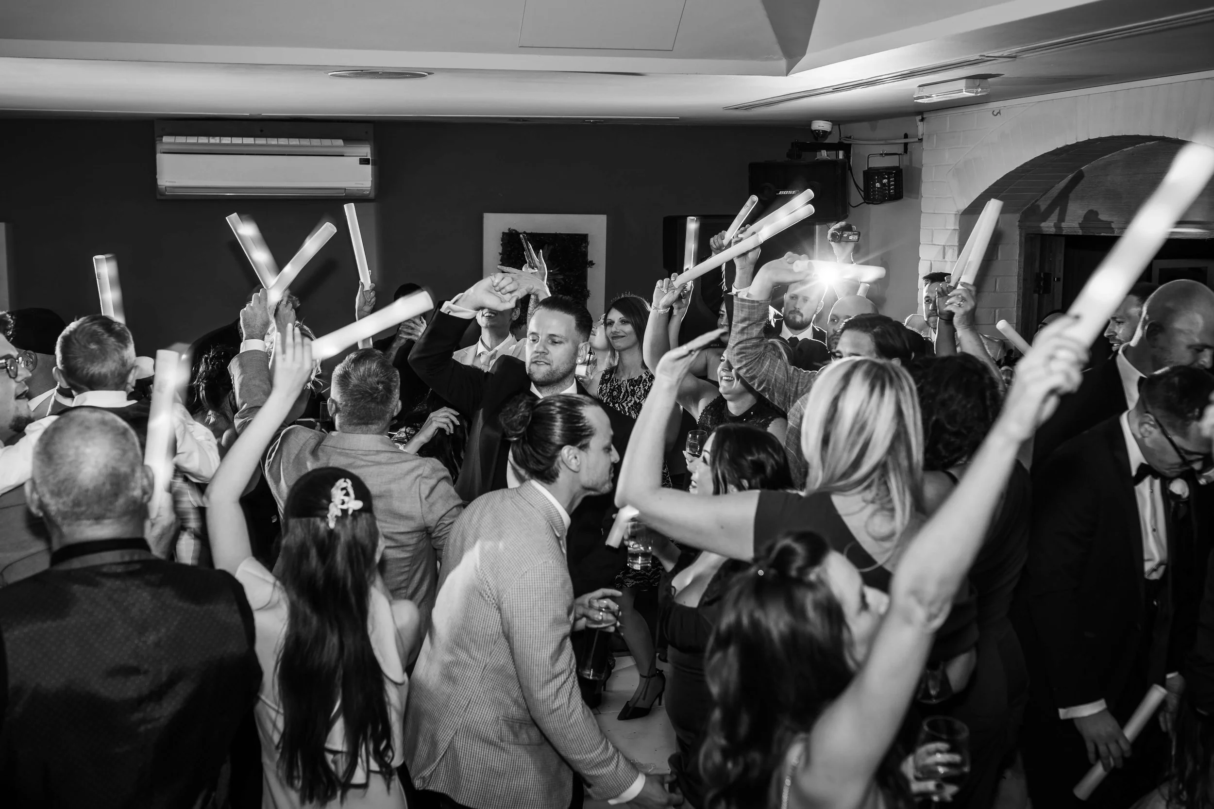 People dancing at a celebration, holding glow sticks in a crowded indoor event.