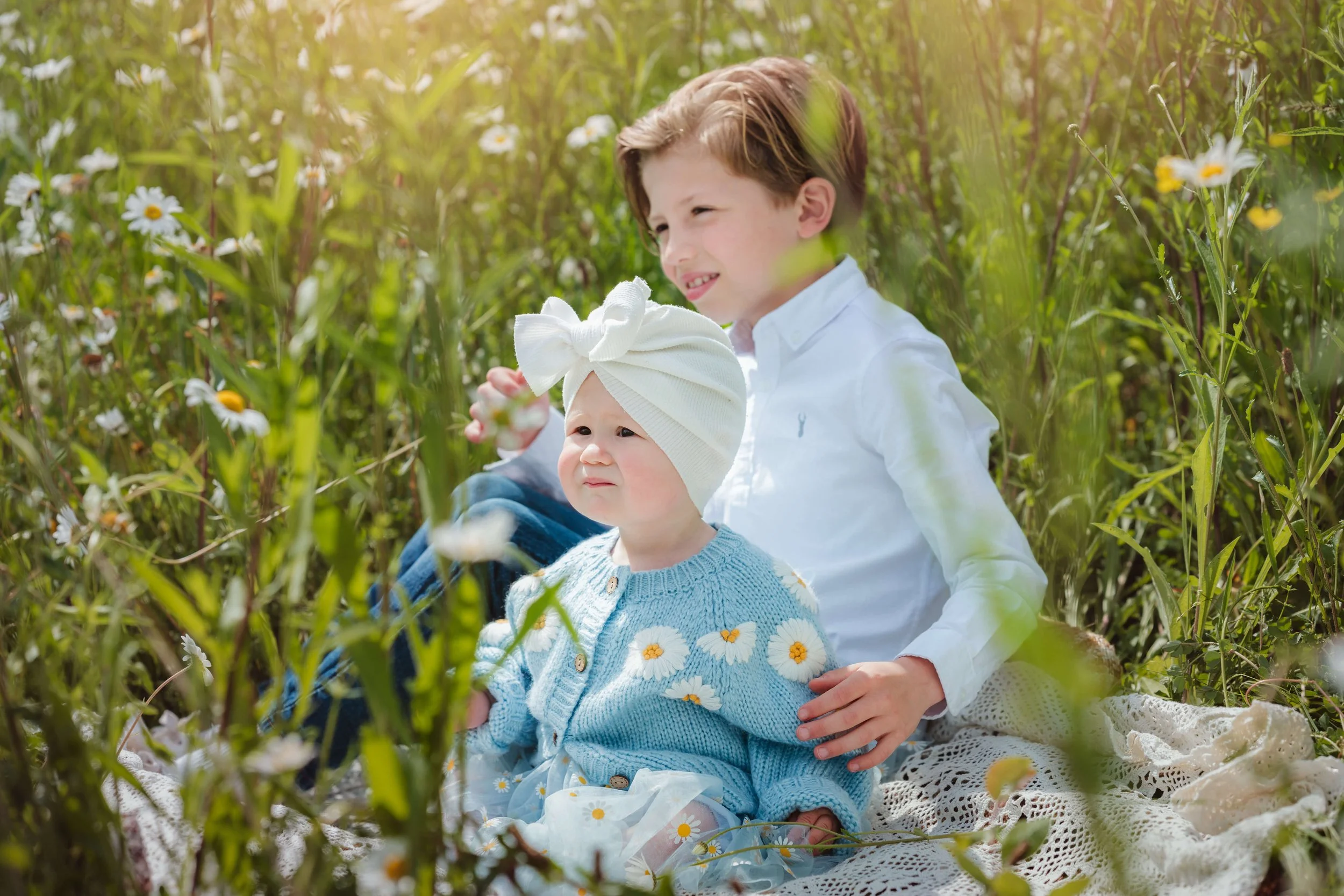 A boy and a baby girl sitting in a field of wildflowers. The boy has short, light brown hair and is wearing a white shirt, while the baby girl is wearing a white hat and a blue sweater with daisies on it.