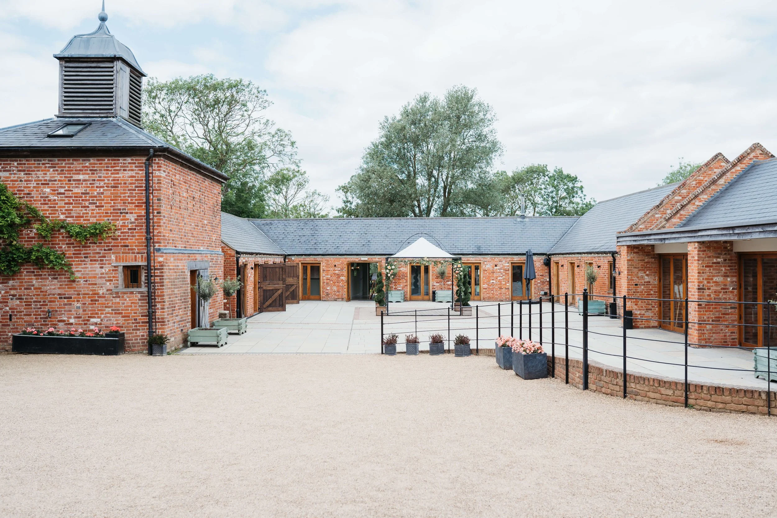 The image shows a courtyard with a gravel area in the foreground, a brick building with a gray roof and wooden doors, and some potted plants and outdoor furniture. Trees are visible in the background under a cloudy sky.