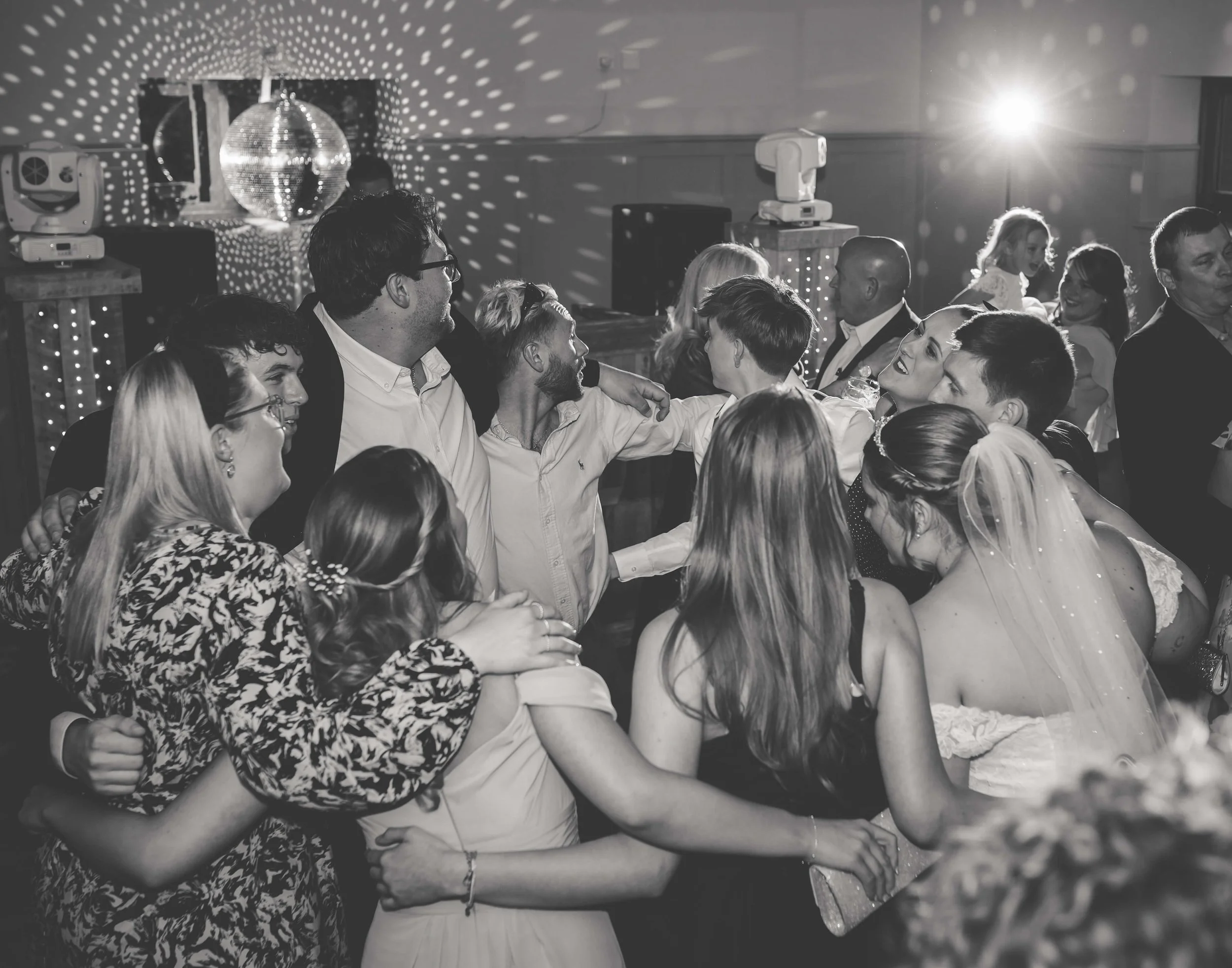 A group of people dancing and celebrating at a party or wedding reception, with a disco ball and stage lights in the background.