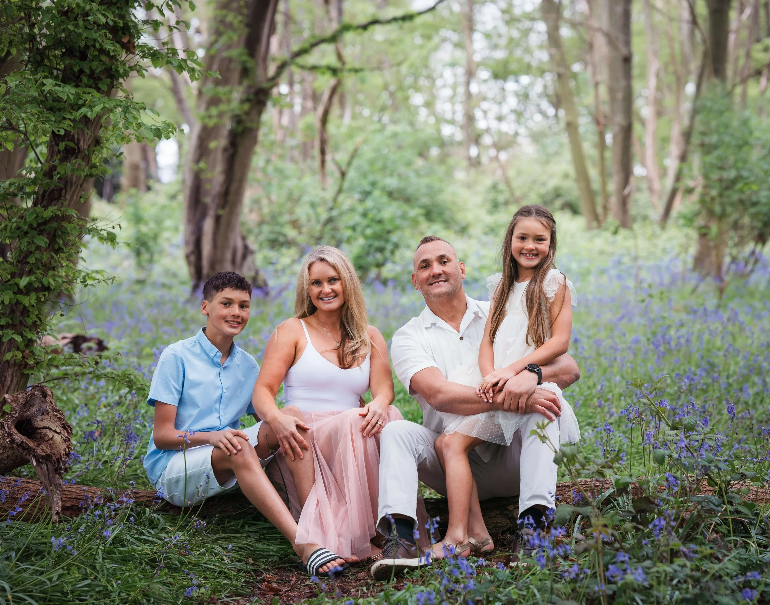 Family of four sitting on a fallen tree in a forest surrounded by blue flowers, smiling at the camera.