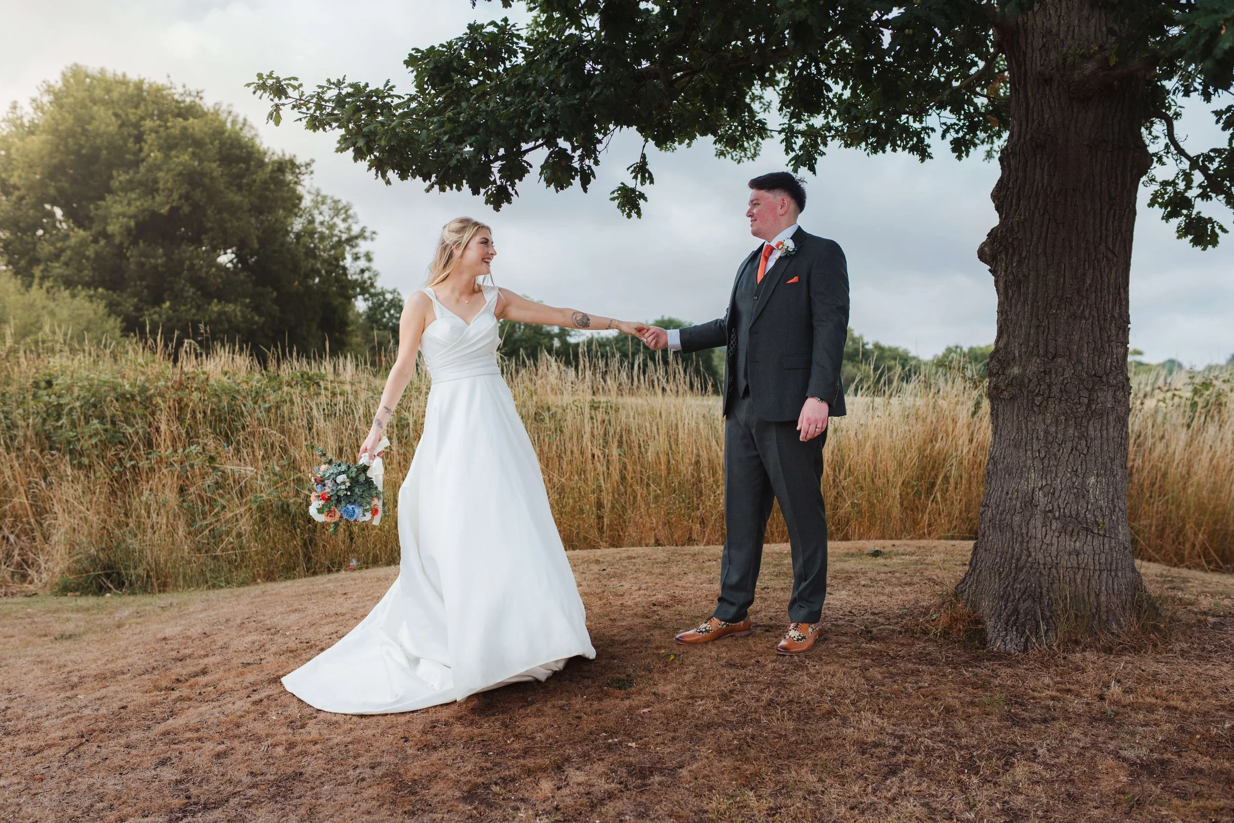 A bride in a white wedding dress holding a bouquet, smiling at a groom dressed in a dark suit, standing outdoors under a large tree, holding hands in a grassy field with a cloudy sky in the background.