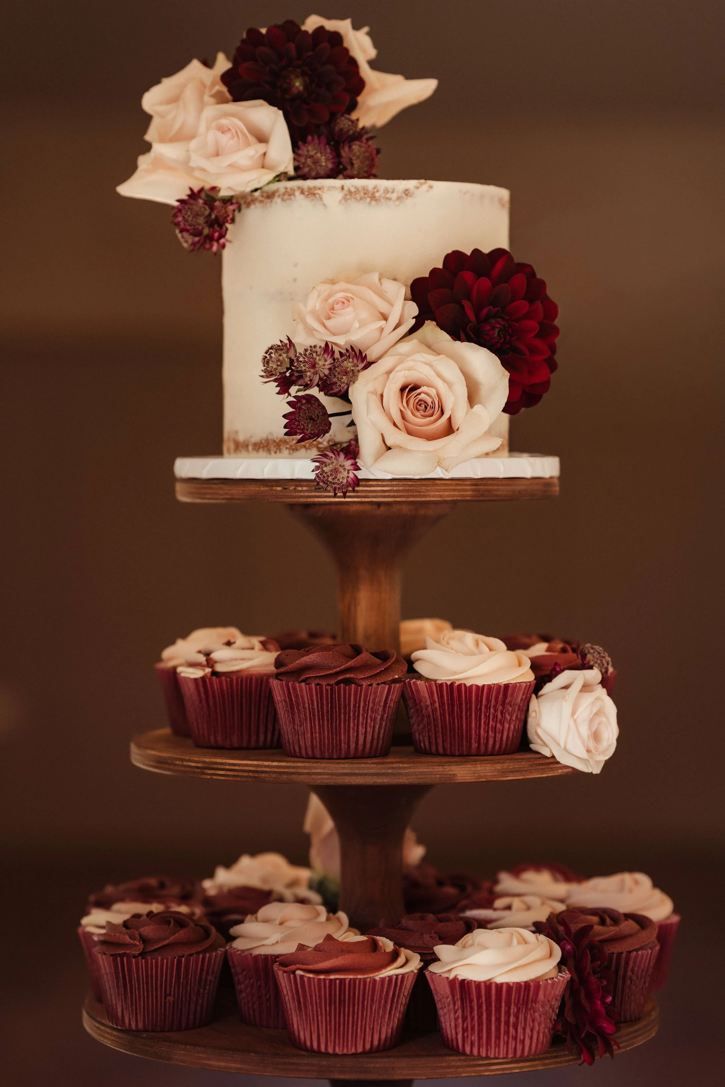 A tiered dessert stand with a white frosted cake decorated with pink and dark red flowers on top and along the side, accompanied by chocolate and vanilla cupcakes with frosting, displayed on a wooden stand against a dark background.