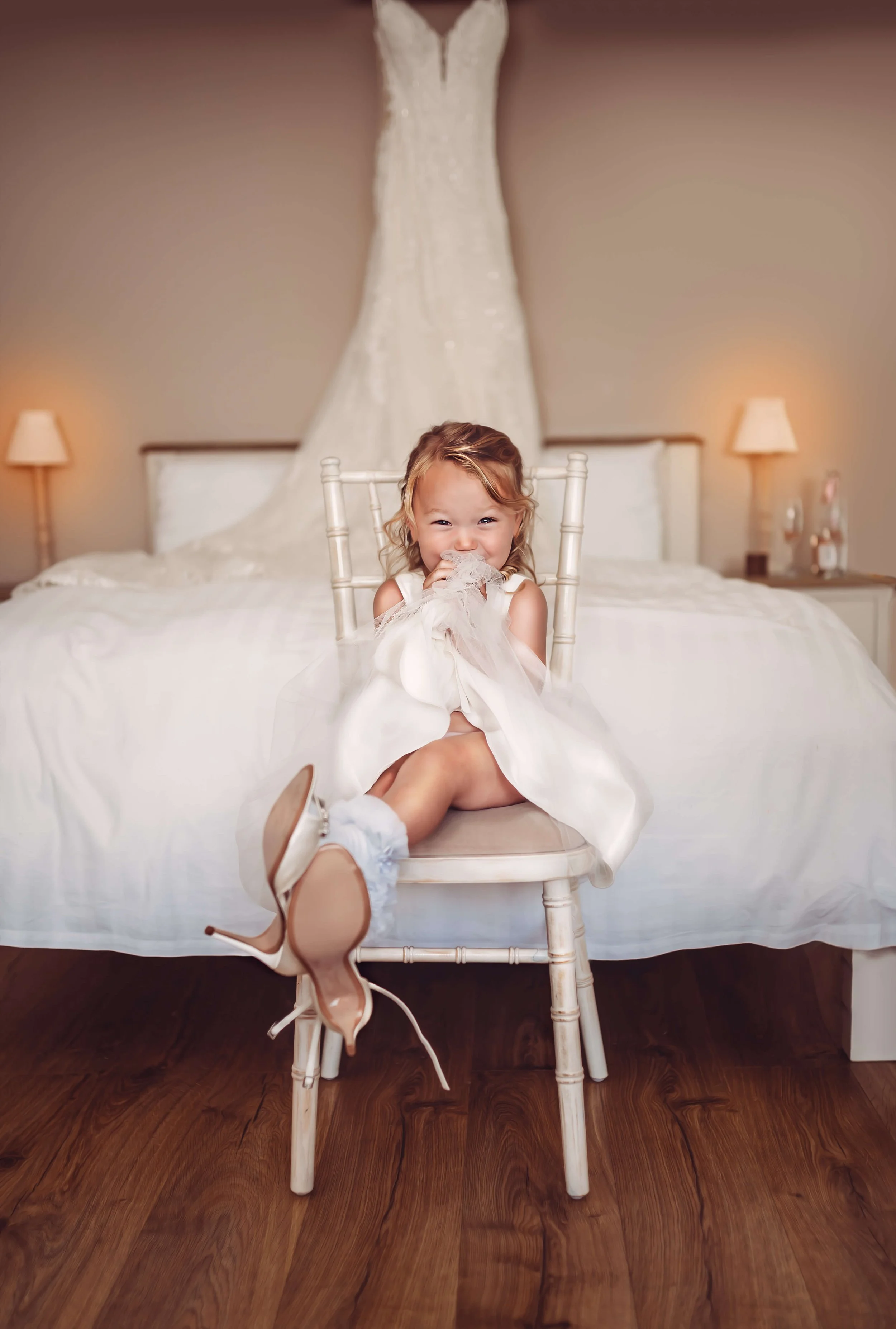 A young girl with curly hair in a white dress sitting on a chair in front of a bed with a wedding dress hanging above it, smiling and holding a piece of fabric near her face.