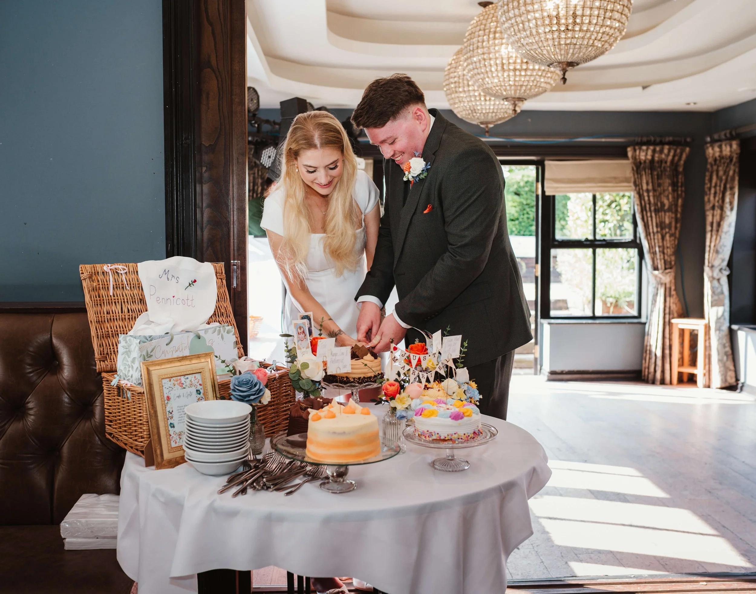 A bride and groom cutting their wedding cake at a celebration, with decorated cakes and gifts on the table.