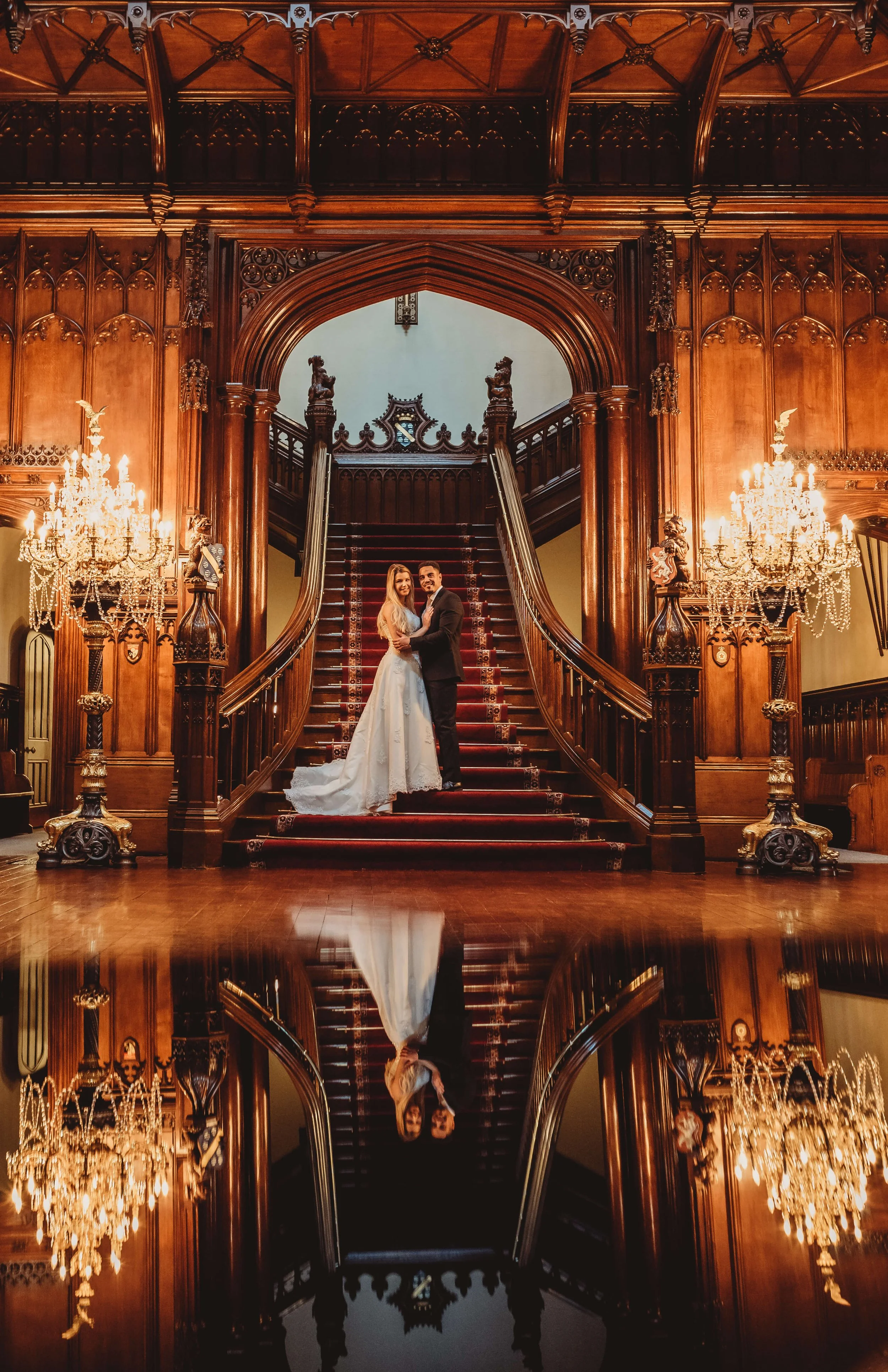 A bride and groom standing on a grand staircase in an ornate wood-paneled hall with chandeliers, reflected in a polished floor.
