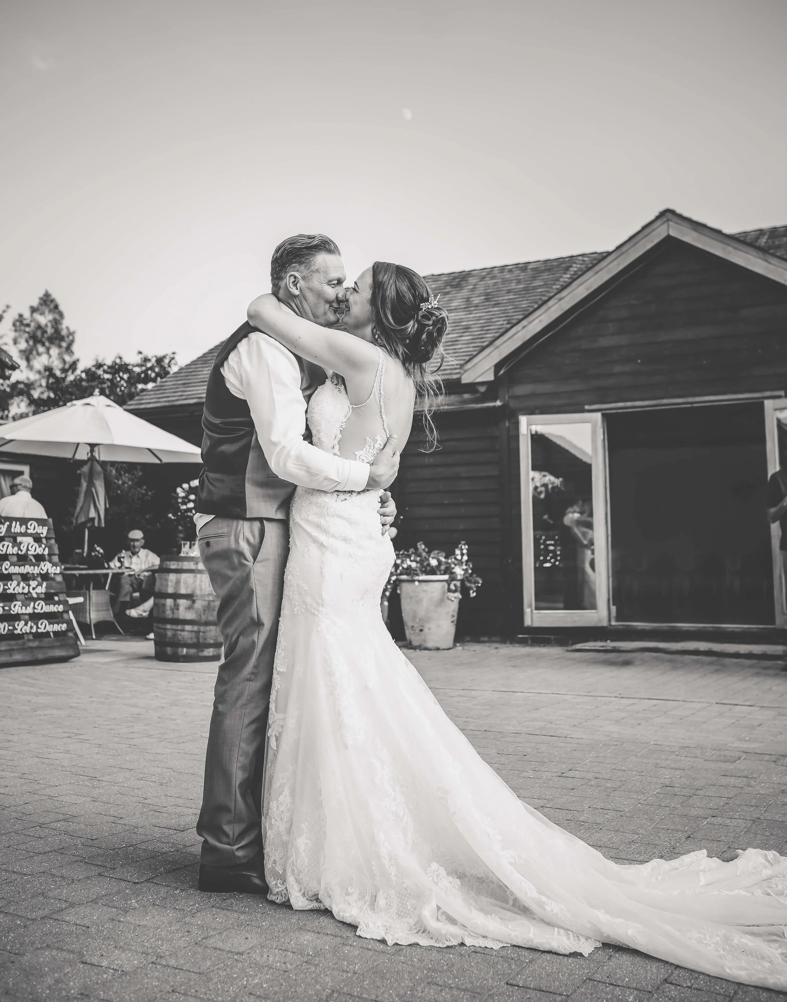 Black and white photo of a bride and groom hugging and kissing outdoors, with a wooden building, potted plants, and a patio with tables and umbrellas in the background.