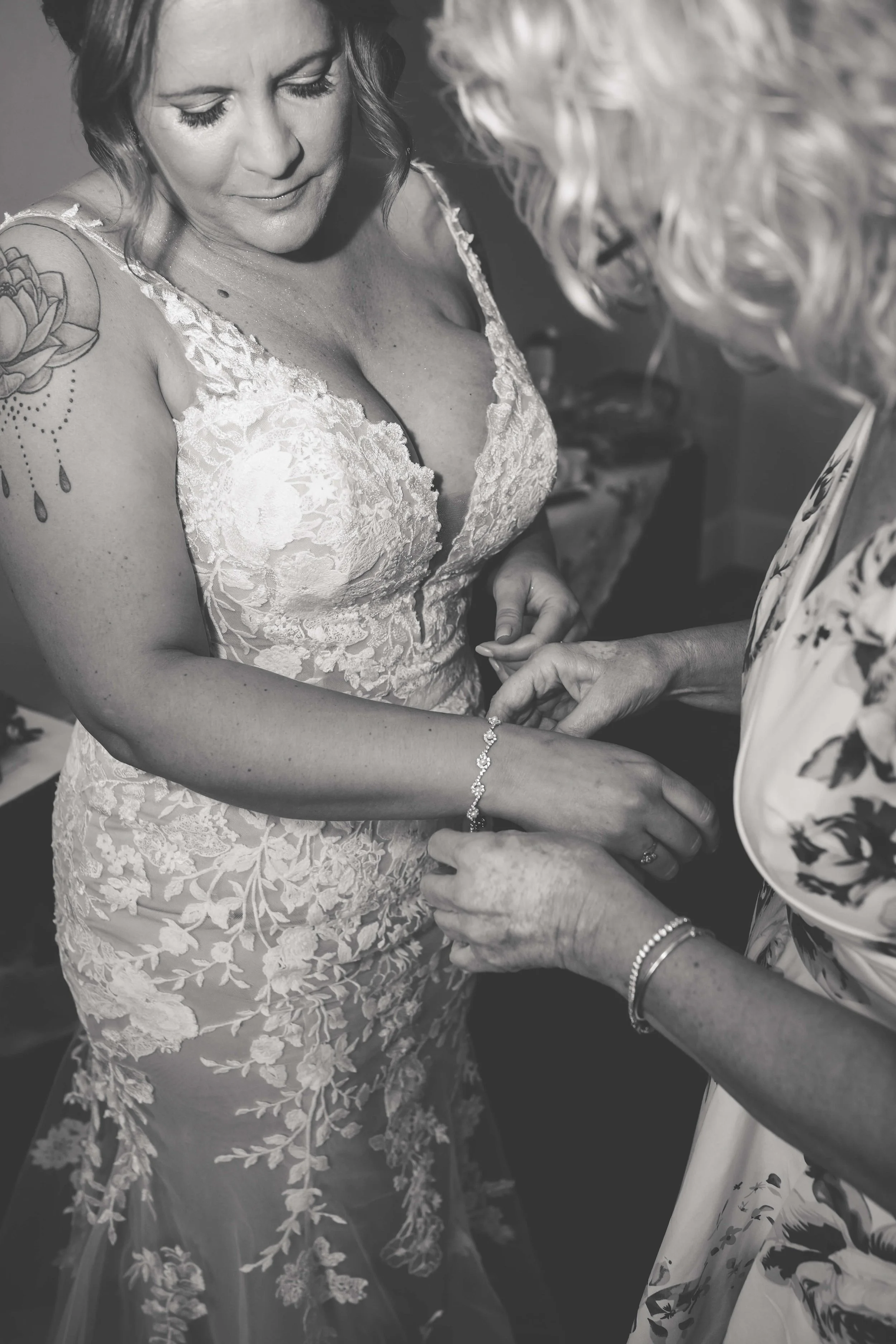 A woman in a lace wedding dress is being helped with jewelry by another woman.