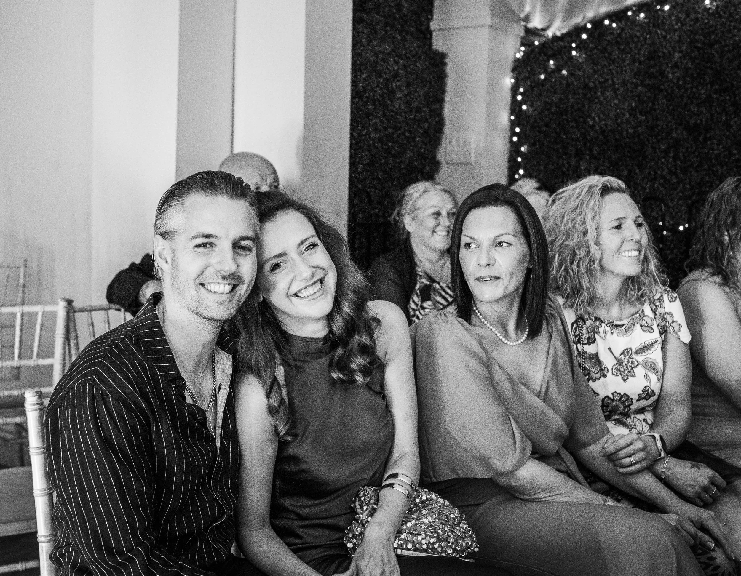 People sitting in a row at an event, smiling and engaging, black and white photo.