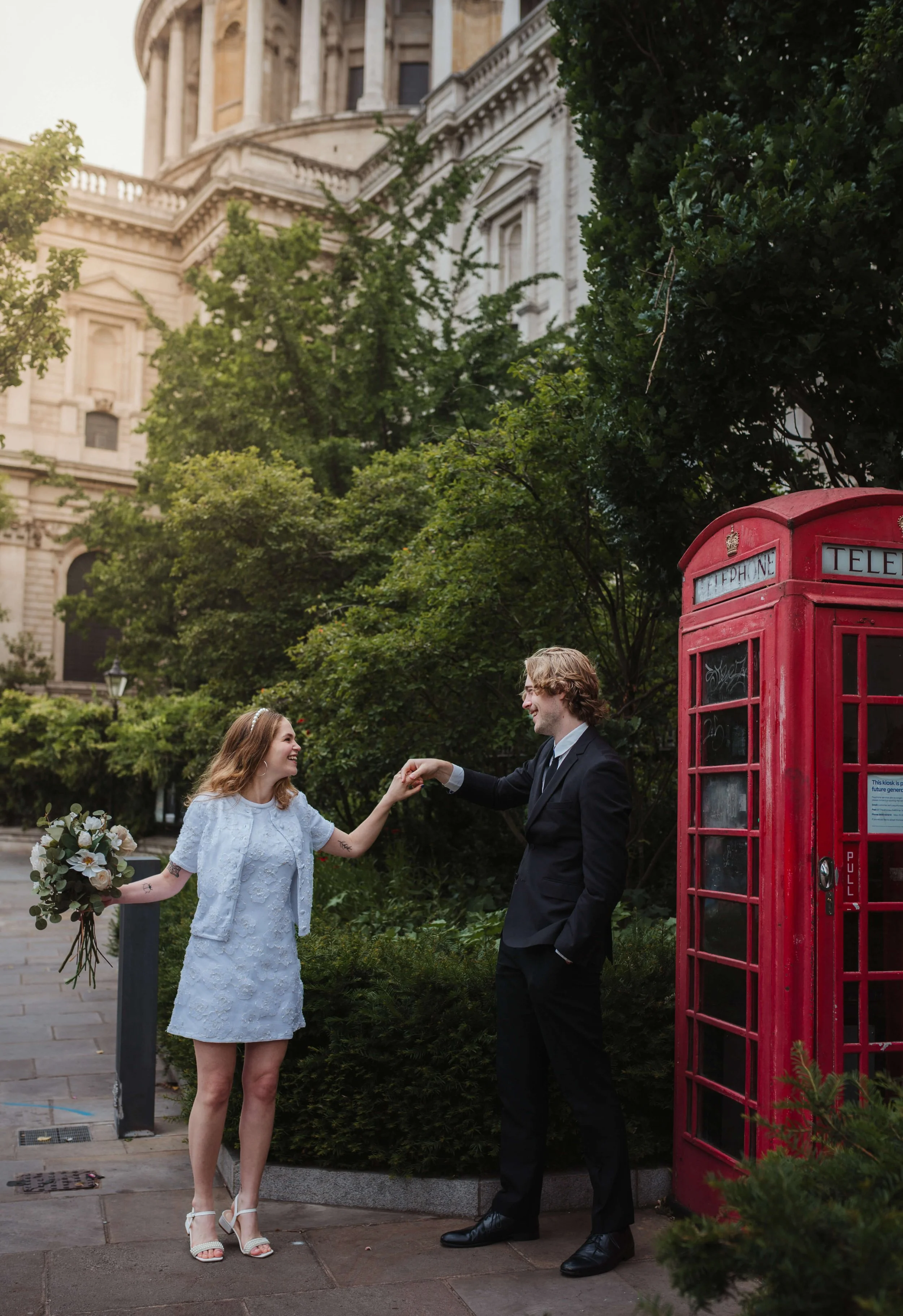 A woman in a white dress and a man in a suit are holding hands and smiling near a red telephone booth on a city street, with a historic building and green trees in the background.