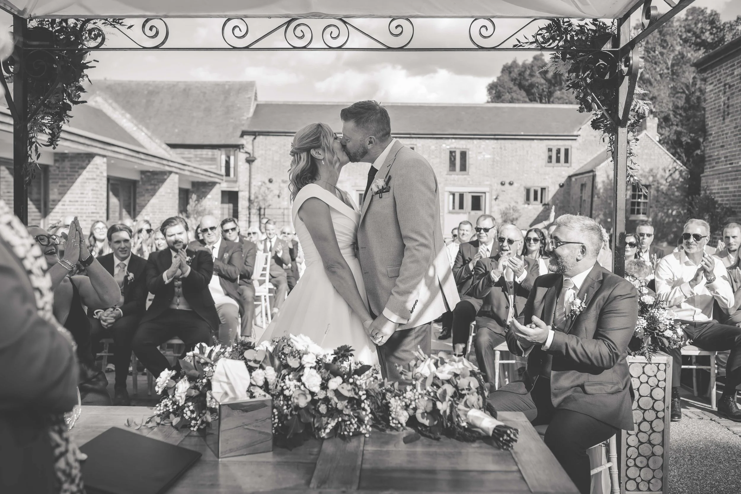 A black and white photo of a wedding ceremony with a couple kissing under an outdoor arch, surrounded by seated guests applauding, with floral arrangements on a table in the foreground.