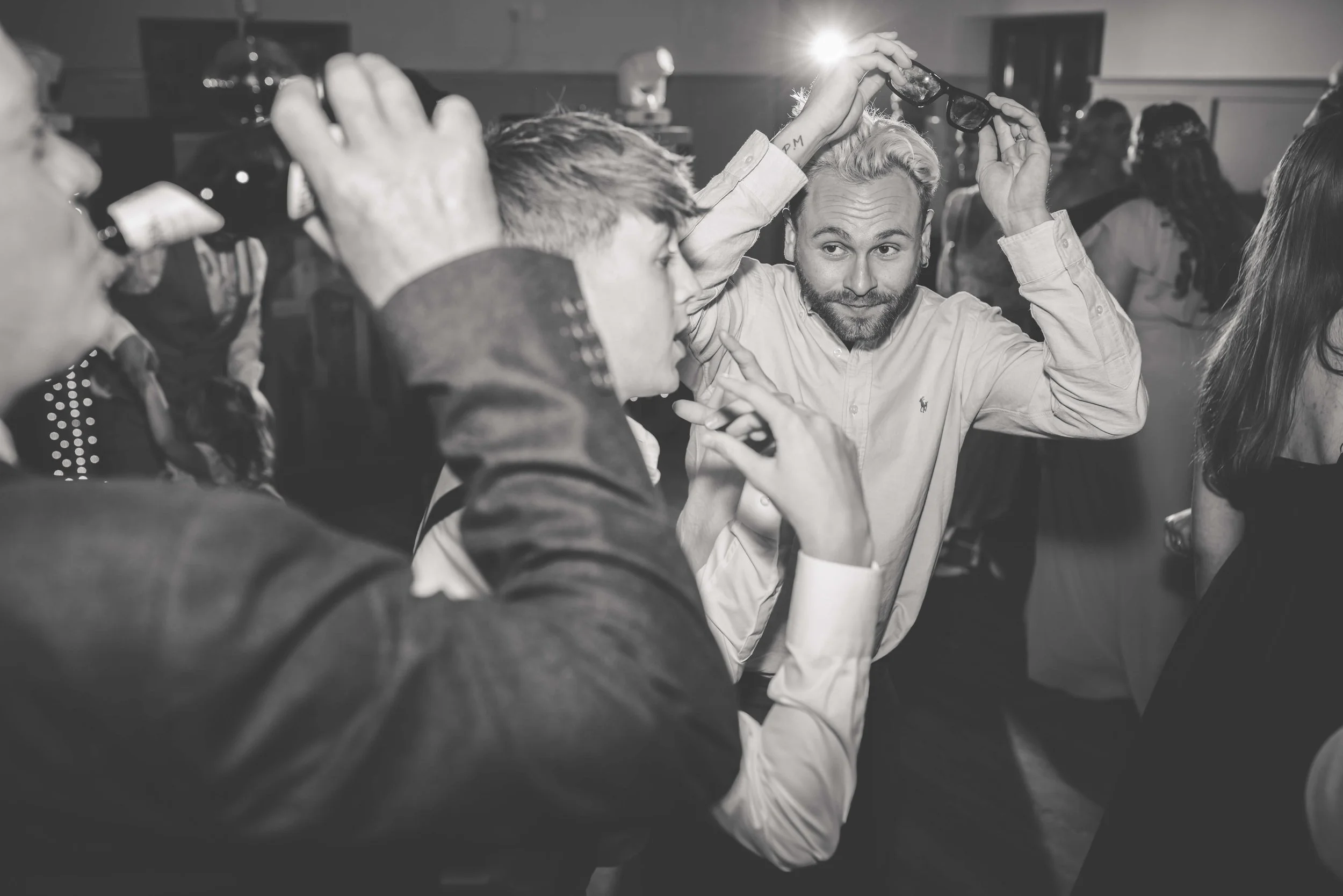People dancing and talking at a party in a crowded room, some with glasses and one person with sunglasses on their head.