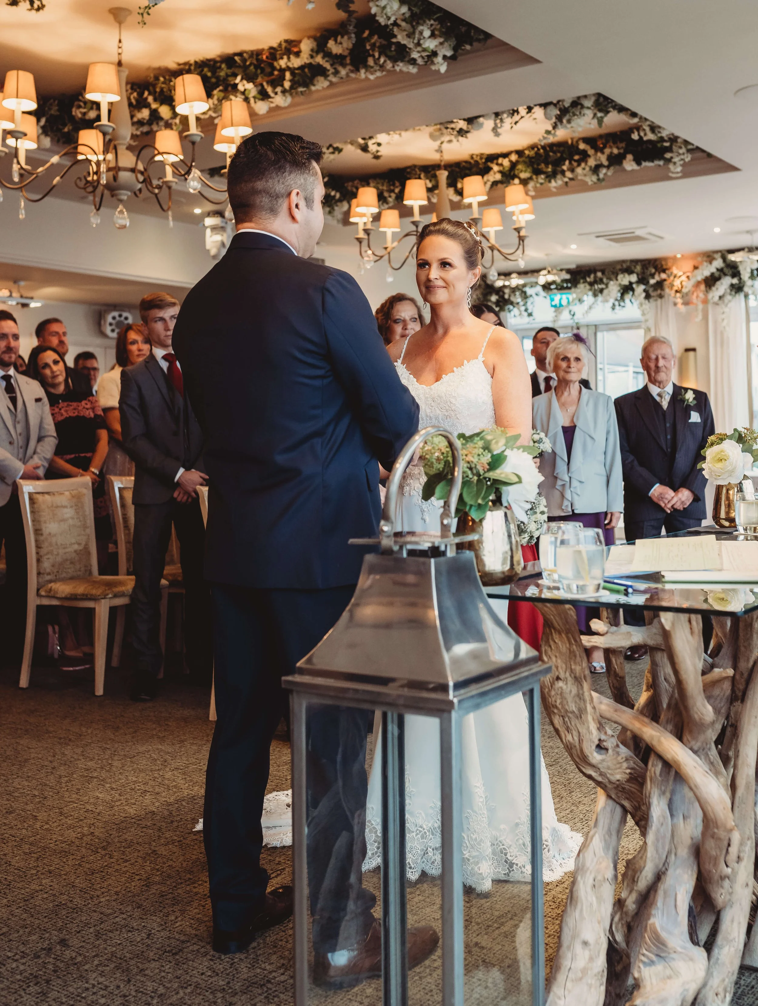 A bride and groom exchanging vows during their wedding ceremony, with guests standing behind them in a decorated indoor venue with floral arrangements and chandeliers.