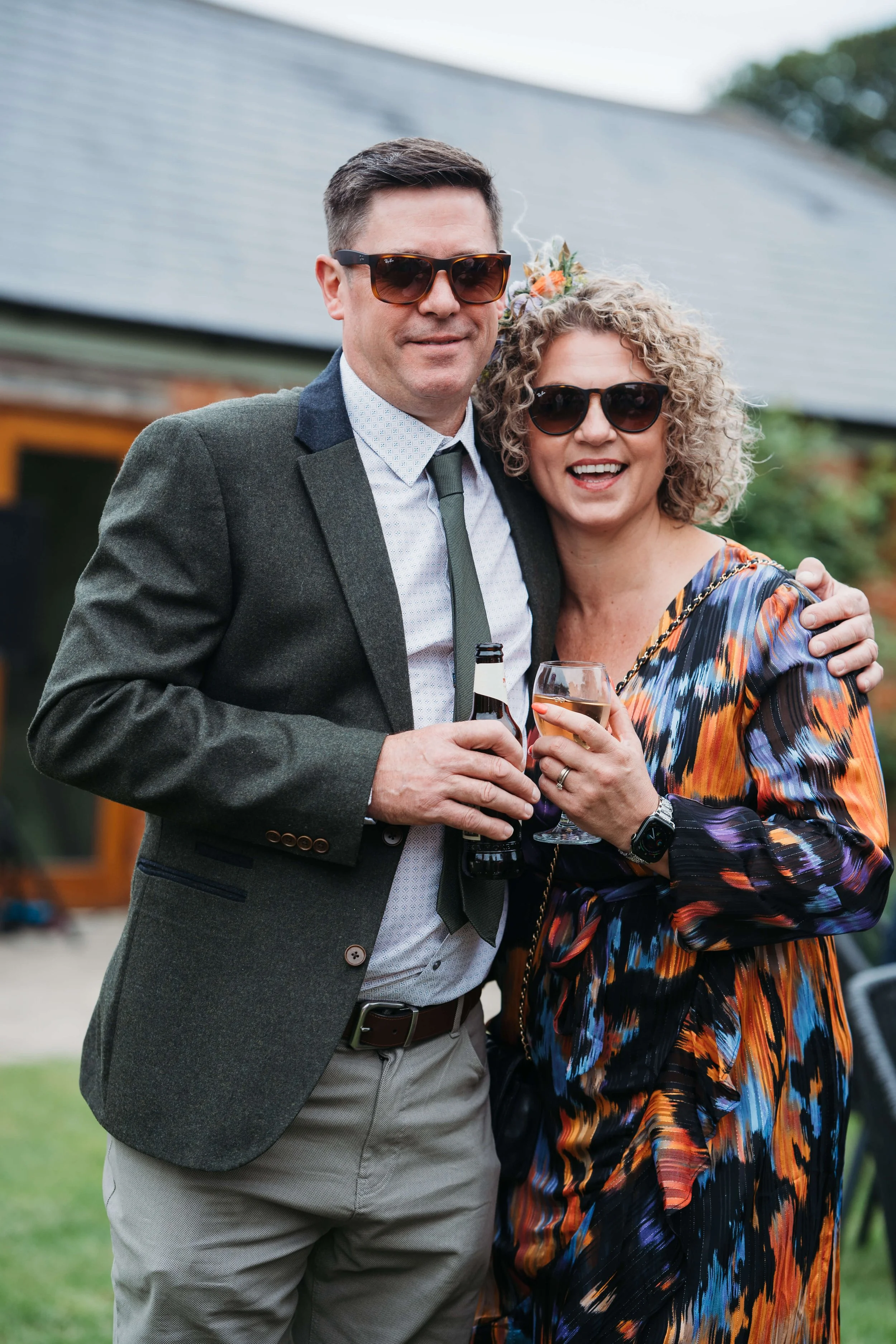 A man and woman are standing outside, hugging, and smiling at a celebration. They are wearing sunglasses, and the woman is holding a glass of wine while the man holds a beer. They are dressed in semi-formal attire, and the background shows a building