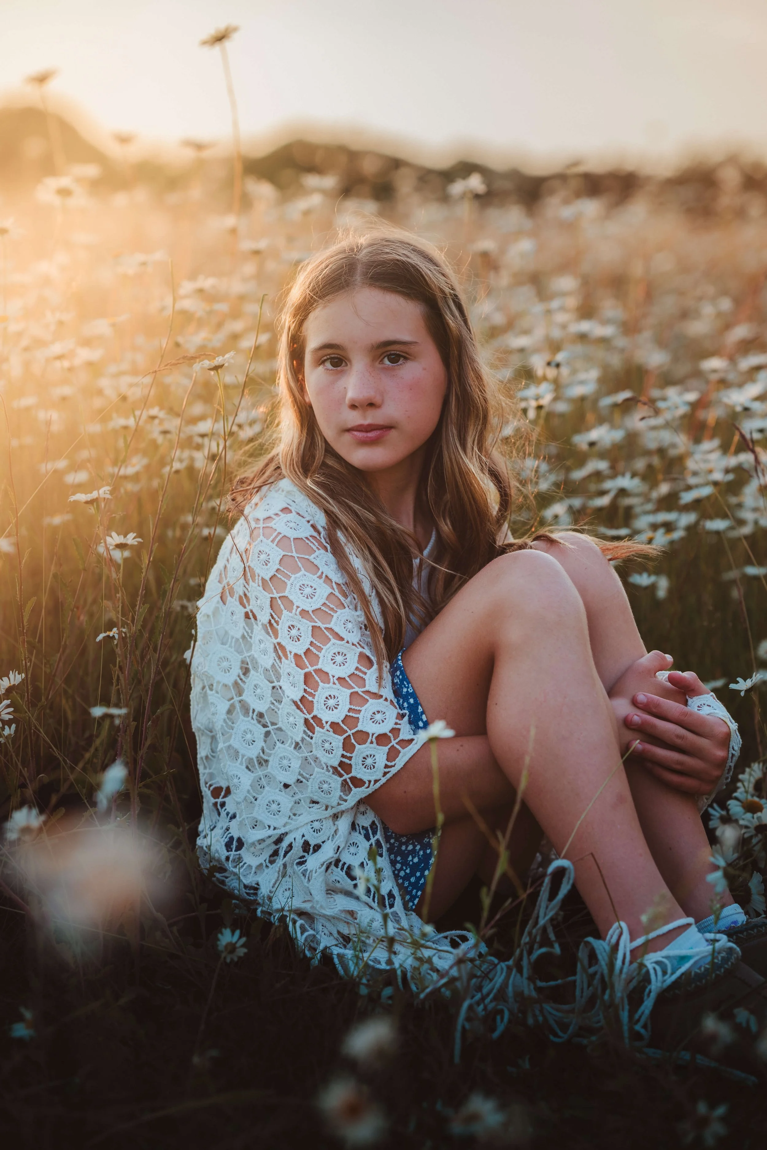 A young girl with long brown hair sitting in a field of daisies during sunset, wearing a white lace top, blue shorts, and sneakers.