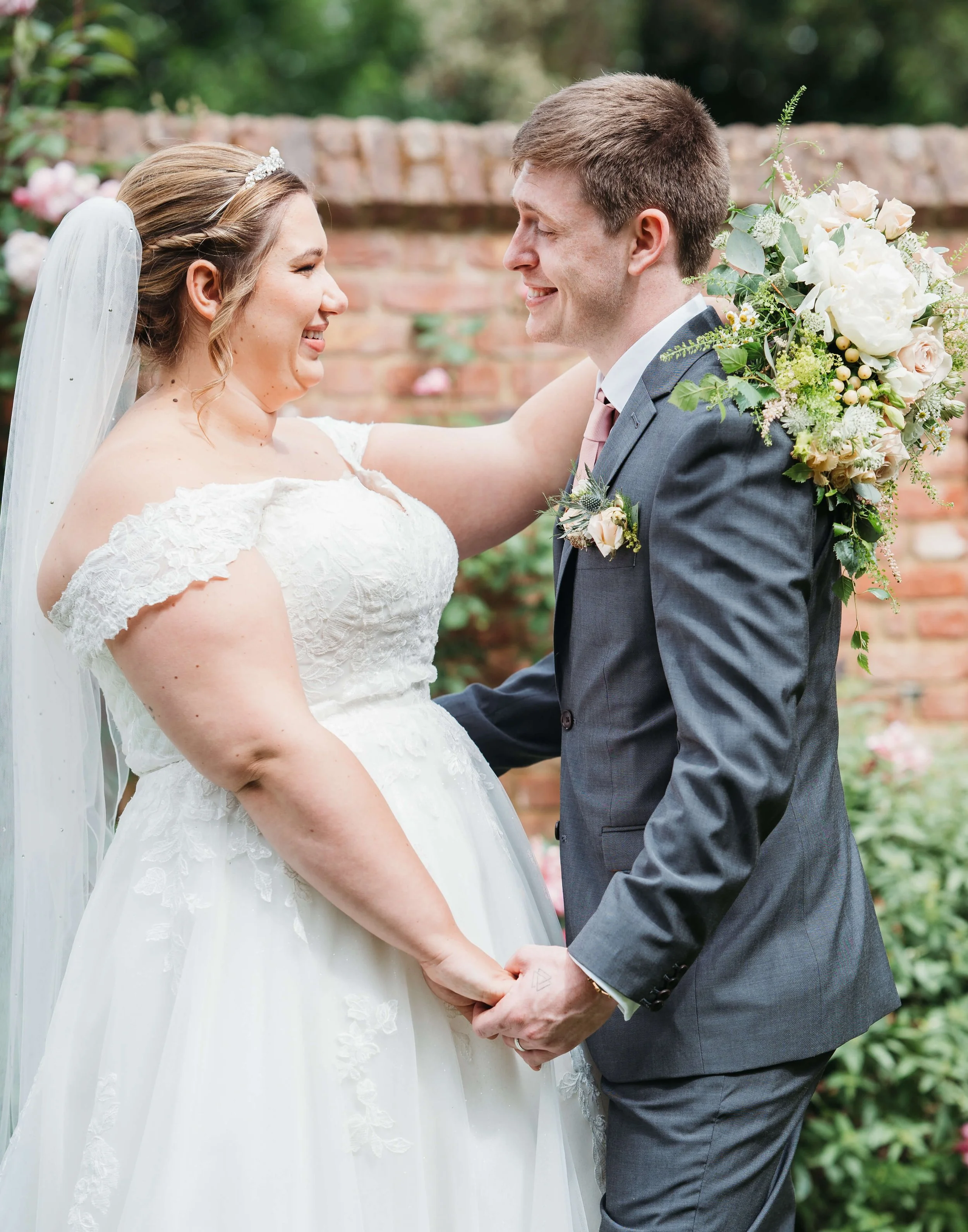 A bride and groom holding hands and smiling at each other outdoors, with the bride wearing a white wedding gown and veil, and the groom in a gray suit with a floral boutonniere and bouquet.