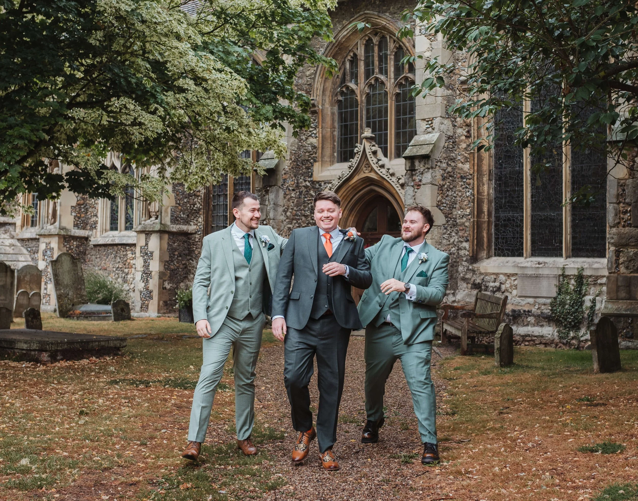 Three men in suits walking and laughing outside a historic stone church with stained glass windows, surrounded by trees and gravestones.