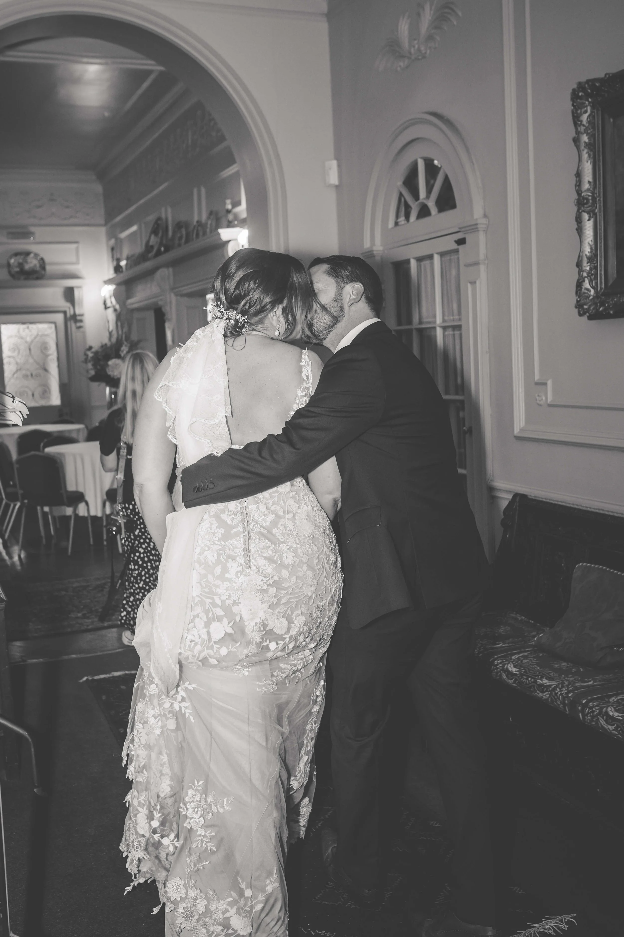 A black-and-white photo of a bride and groom sharing a kiss at their wedding reception, with the groom kneeling and the bride in a lace wedding dress and veil.