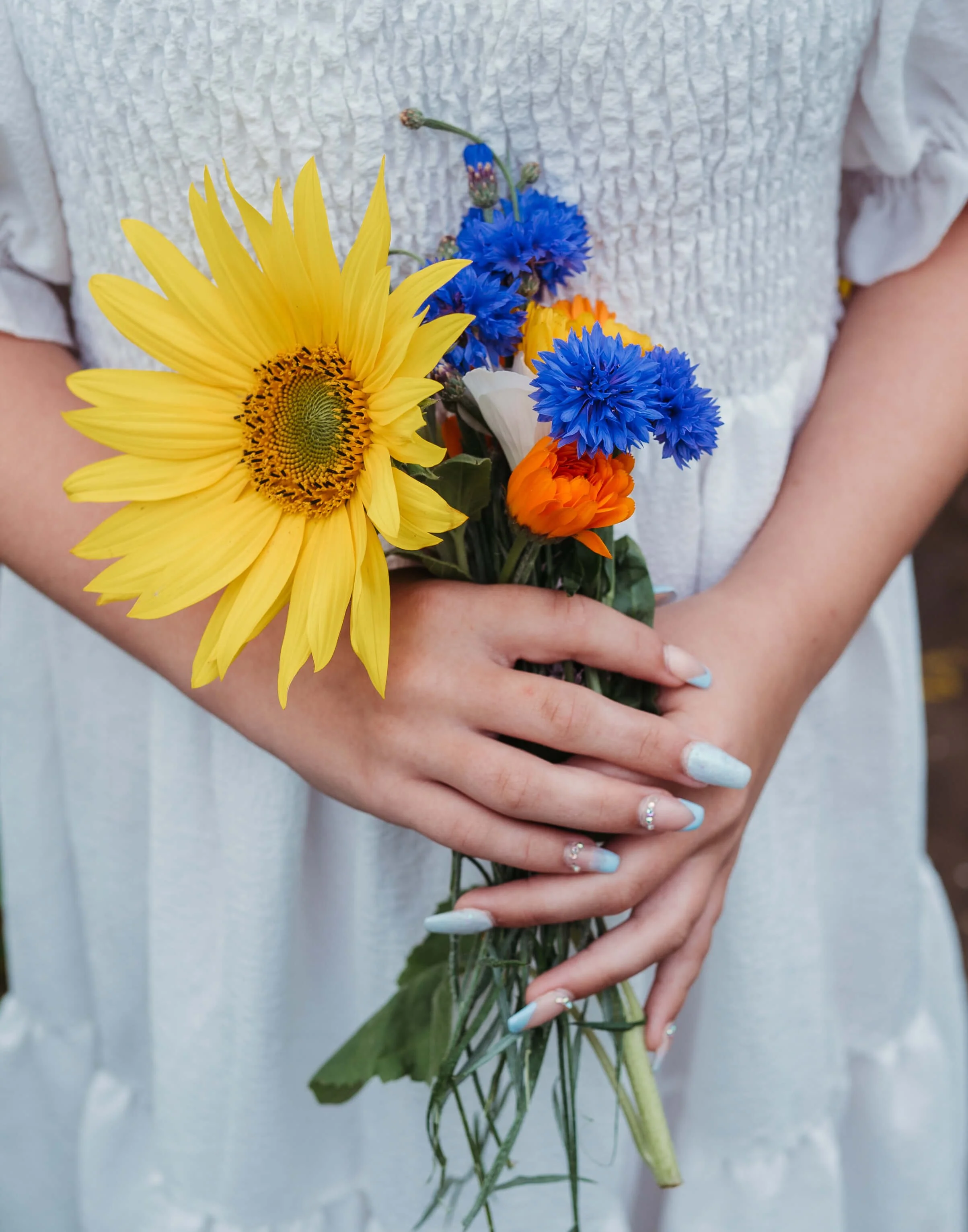 Person holding a bouquet of colorful flowers including a large yellow sunflower, blue cornflowers, orange marigolds, and white lilies.