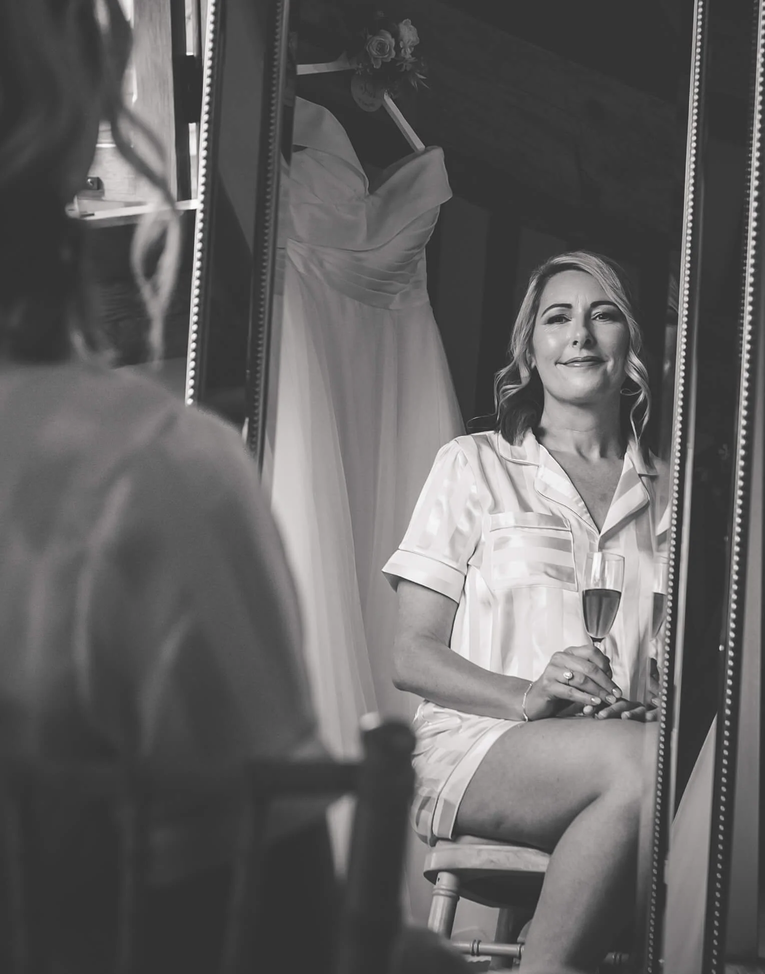 A woman sitting on a stool, holding a glass of champagne, looking into a mirror. A wedding dress is hanging in the background.