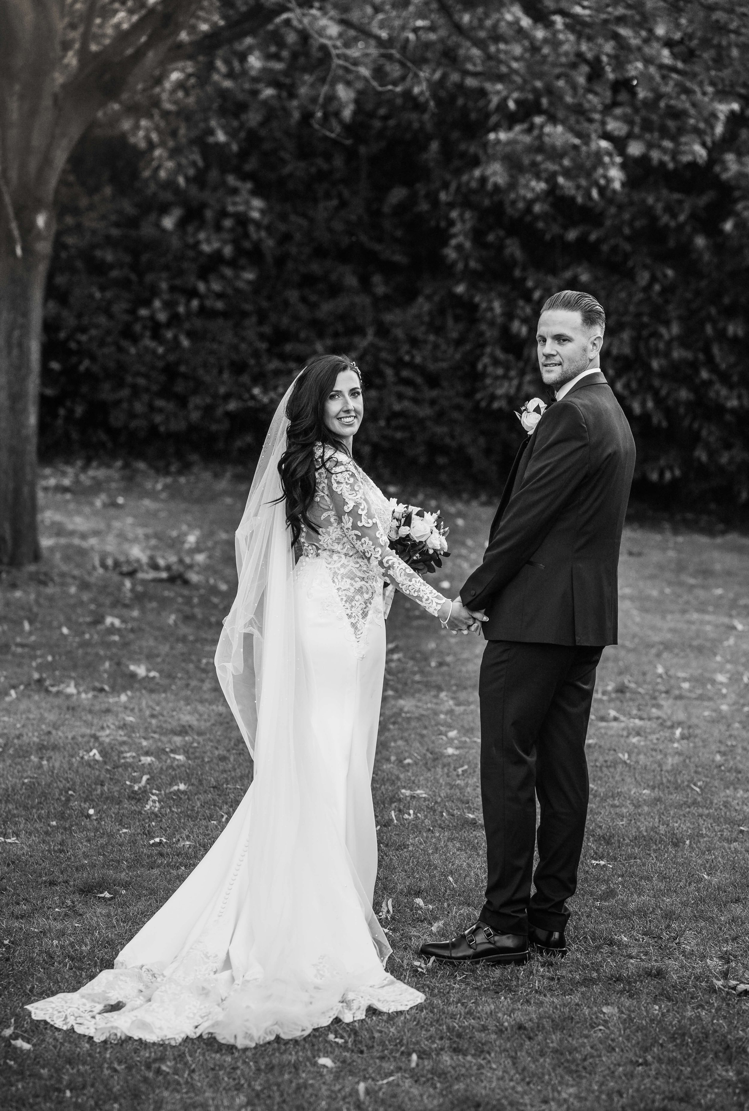 A black and white photograph of a bride and groom holding hands outdoors, standing on grass near a large tree and bushes. The bride is wearing a lace wedding gown with long sleeves and a veil, and the groom is in a dark suit with a boutonniere.