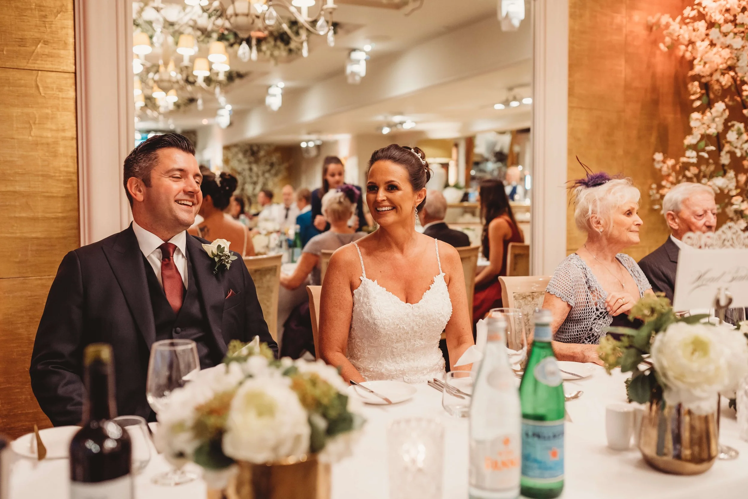 A bride and groom sitting at a wedding reception table, smiling and looking at each other. The bride wears a white lace wedding dress and the groom wears a dark suit with a boutonniere. Other guests are visible in the background, and the table is ado