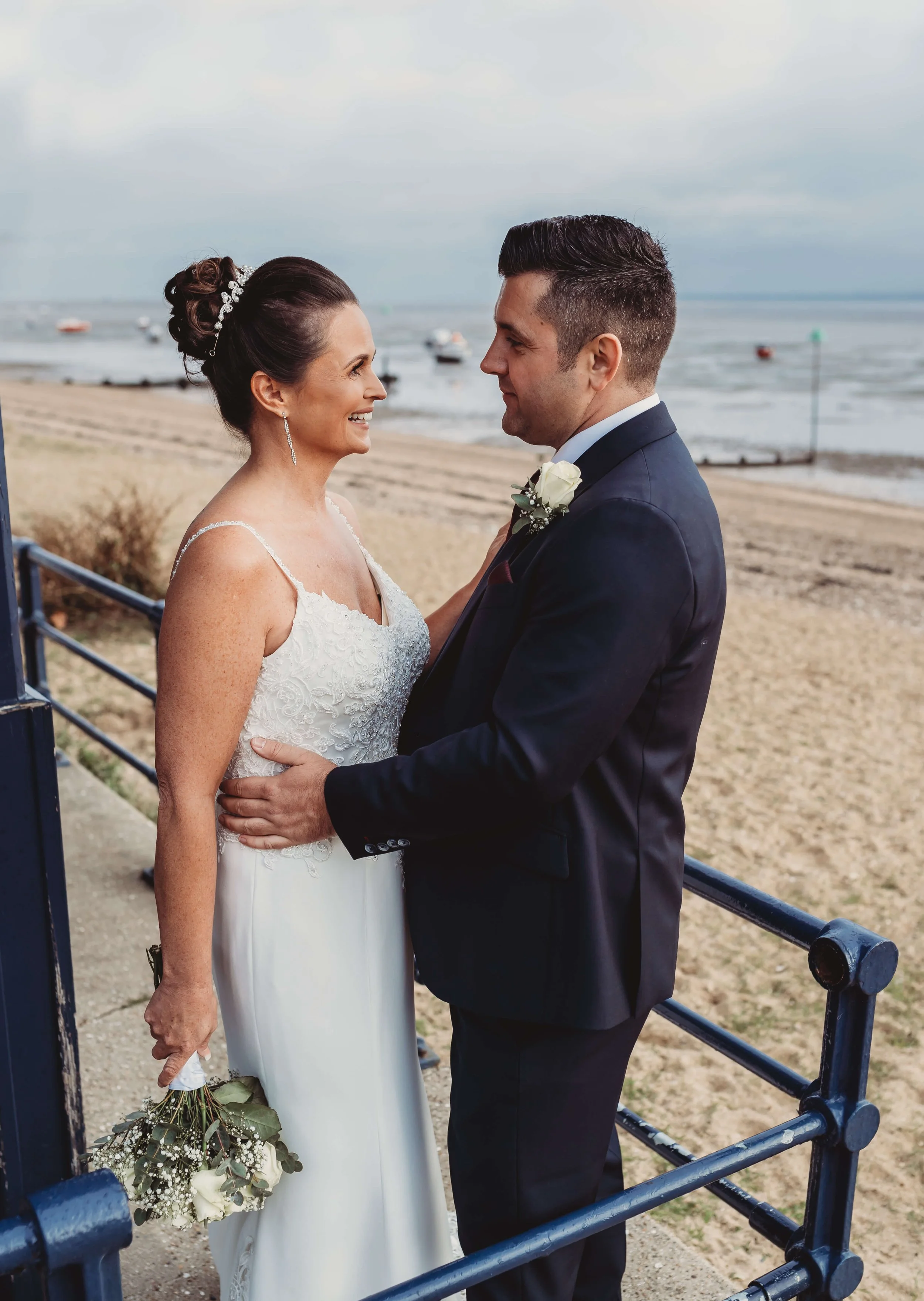A bride and groom standing close together on a beach, smiling at each other, with the bride holding a bouquet of white flowers.