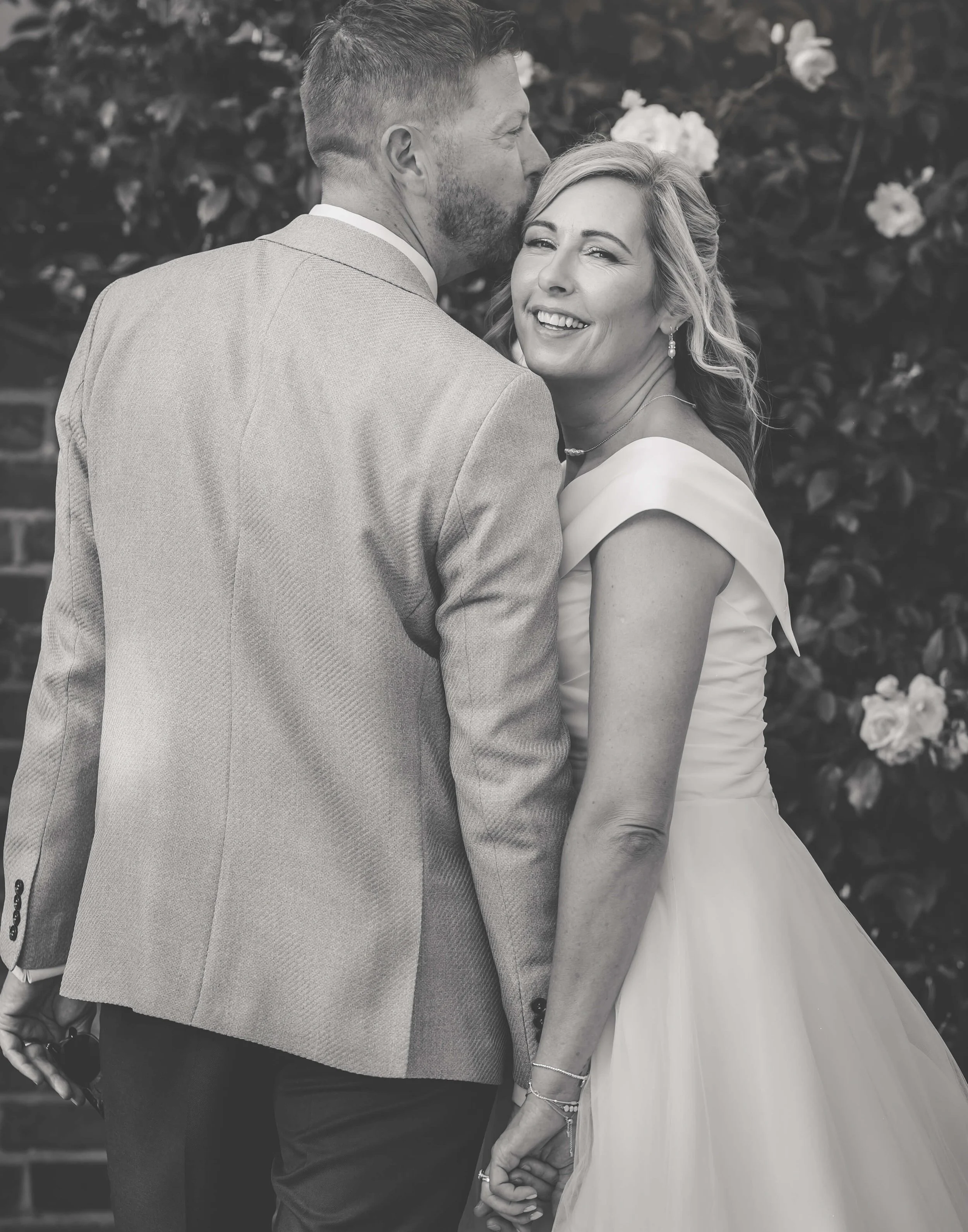 A black-and-white photo of a couple holding hands, with the man whispering into the woman's ear. The woman is smiling and dressed in a wedding gown, while the man is wearing a suit, and background flowers are visible.