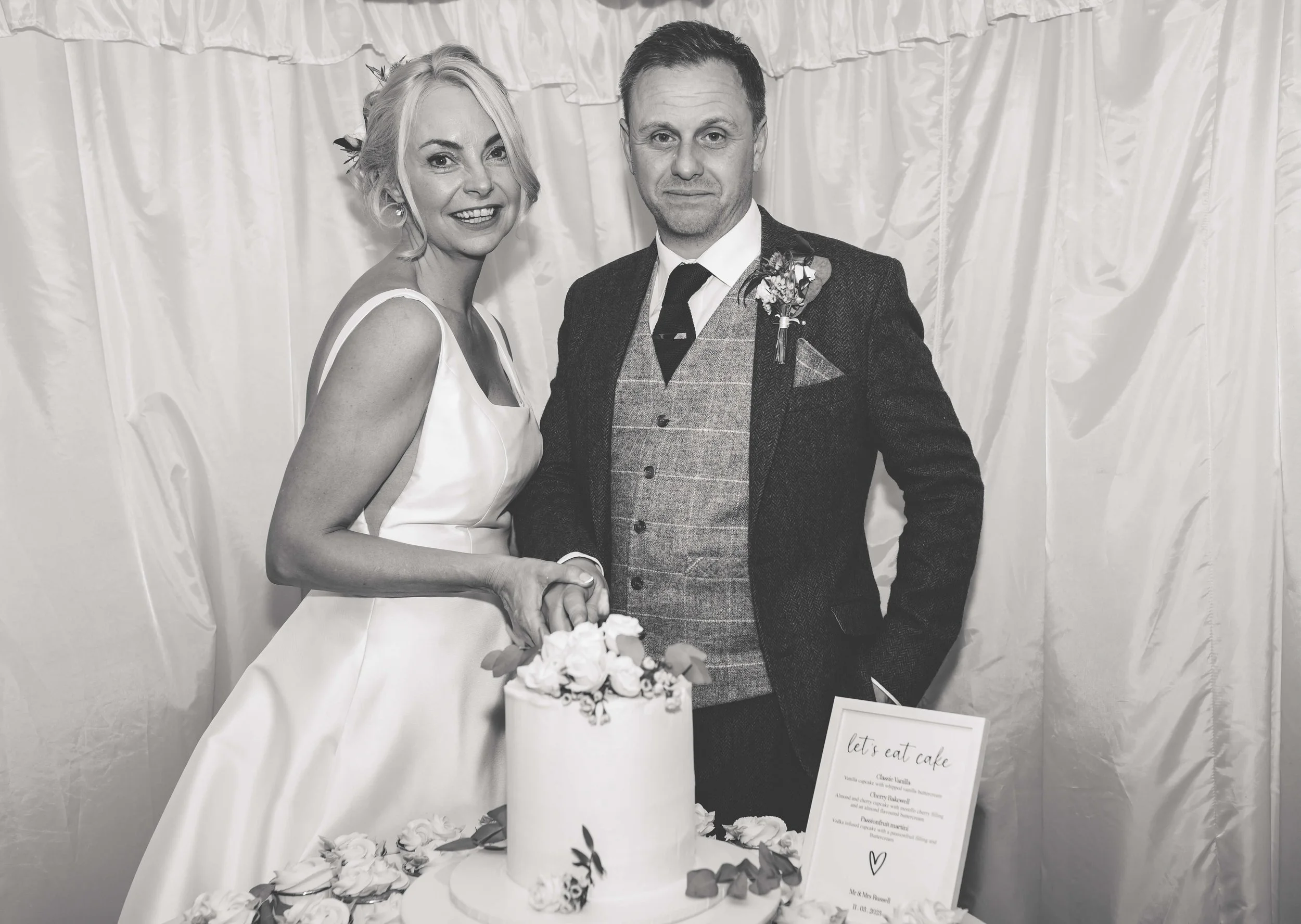 Black and white photo of a bride and groom at their wedding, standing behind a wedding cake, holding hands, with a curtain backdrop.