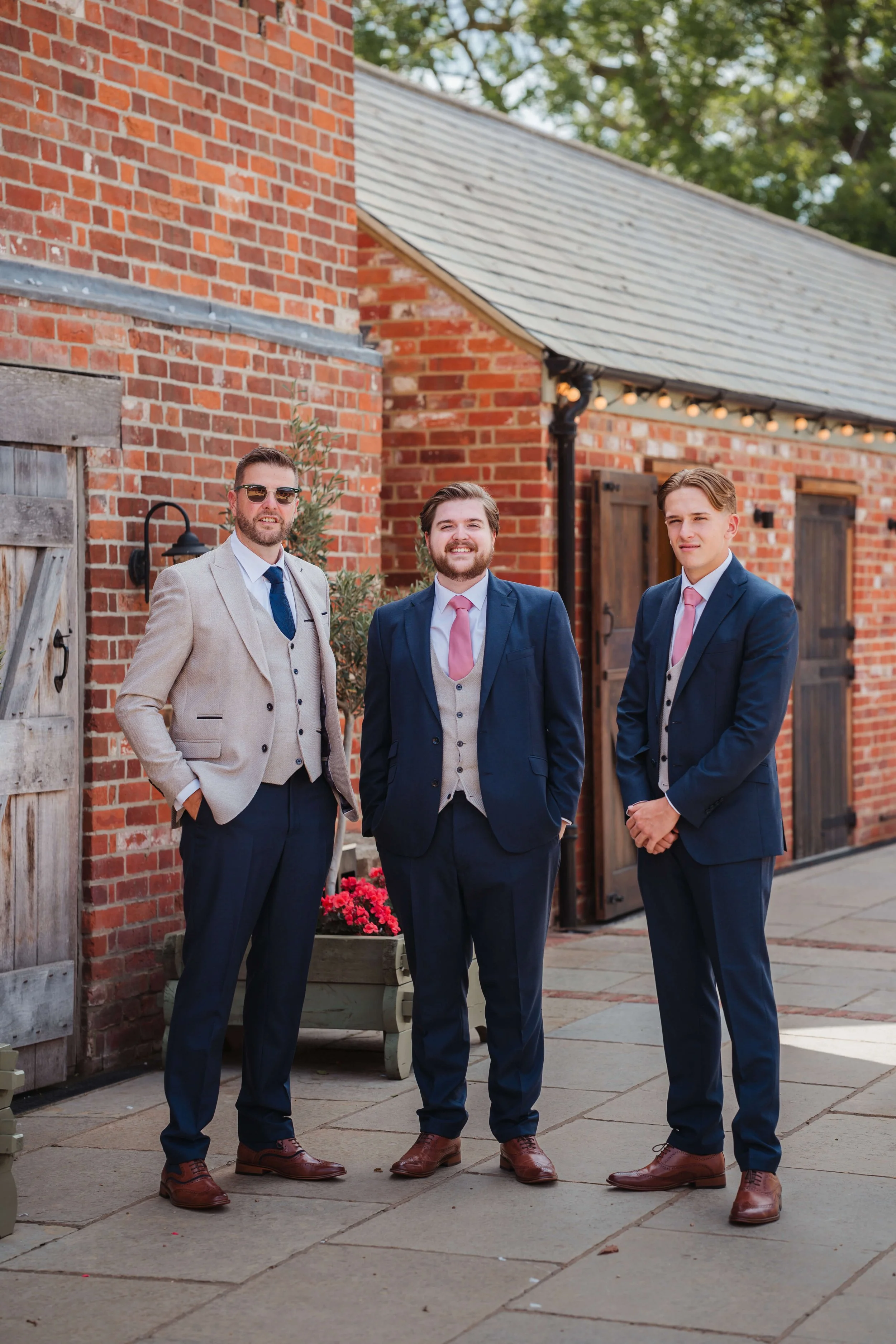 Three men dressed in formal suits standing outdoors near a brick building with wooden doors and string lights.