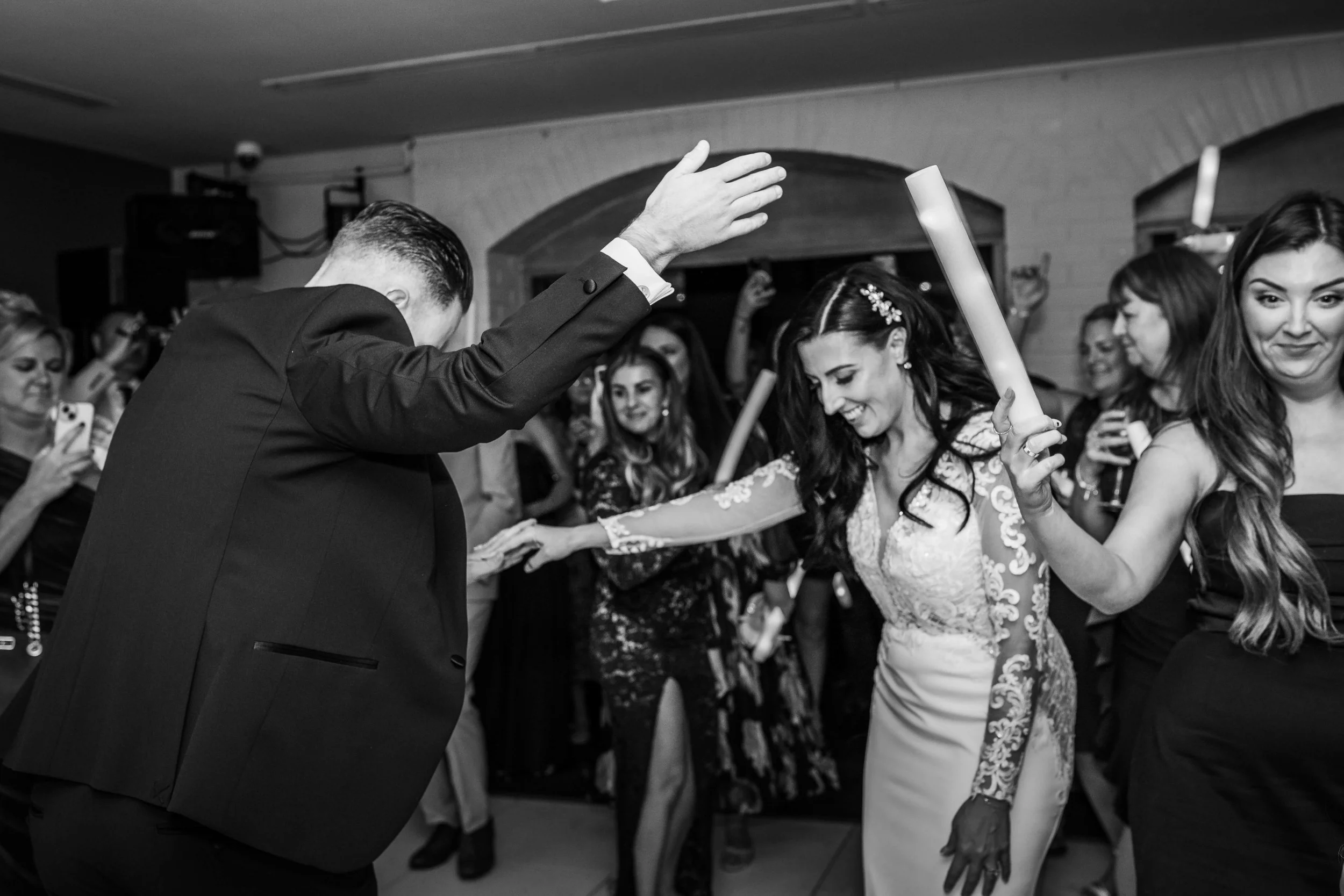 A group of people dancing at a celebration, including a woman in a wedding dress and a man in a suit, with others in the background holding phones and drinks.