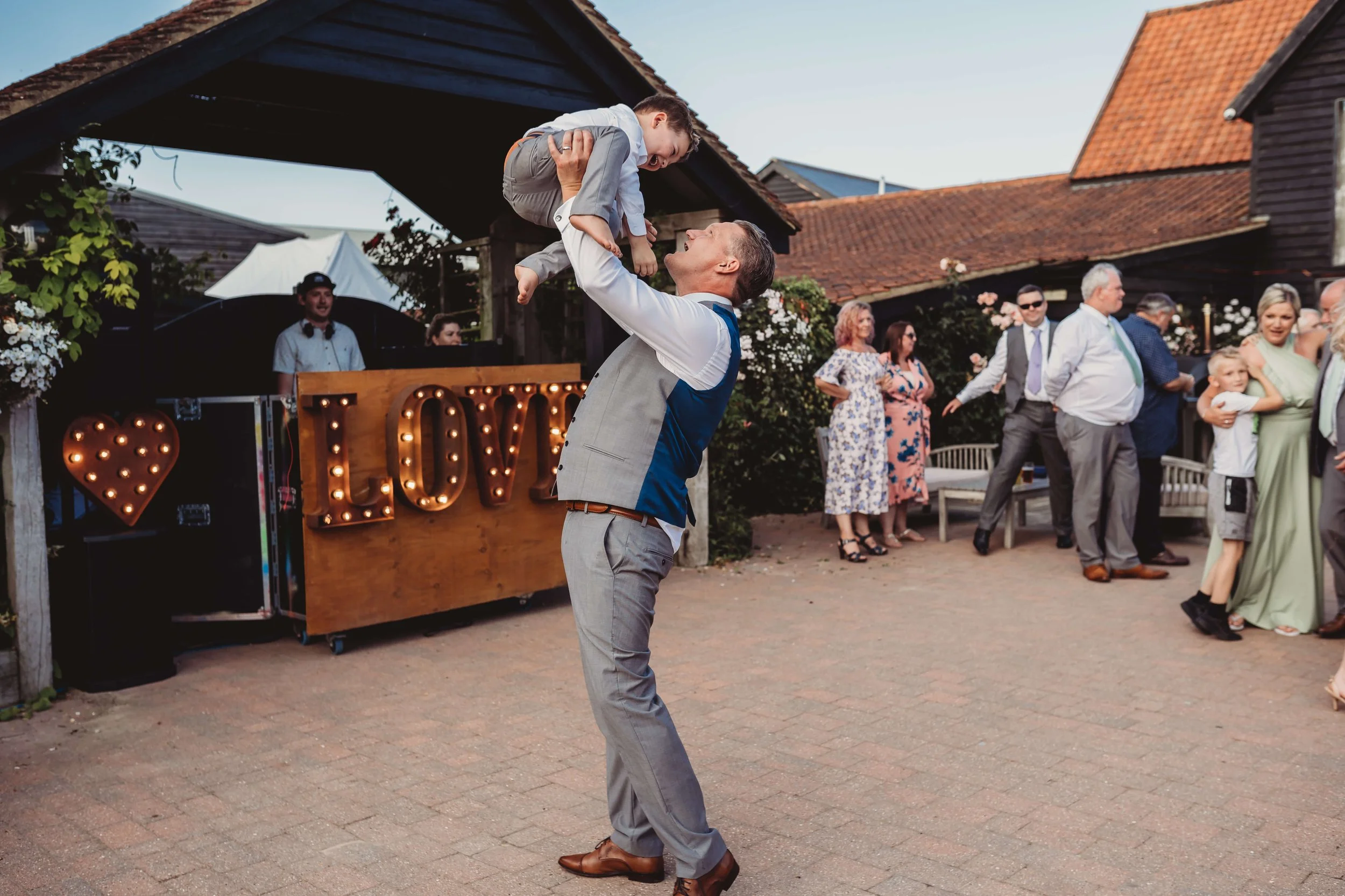 A man in a gray suit and brown shoes is lifting a young boy dressed in a light blue shirt and gray pants during an outdoor celebration, possibly a wedding reception. The background shows a group of people, some dancing and others watching, with a dec