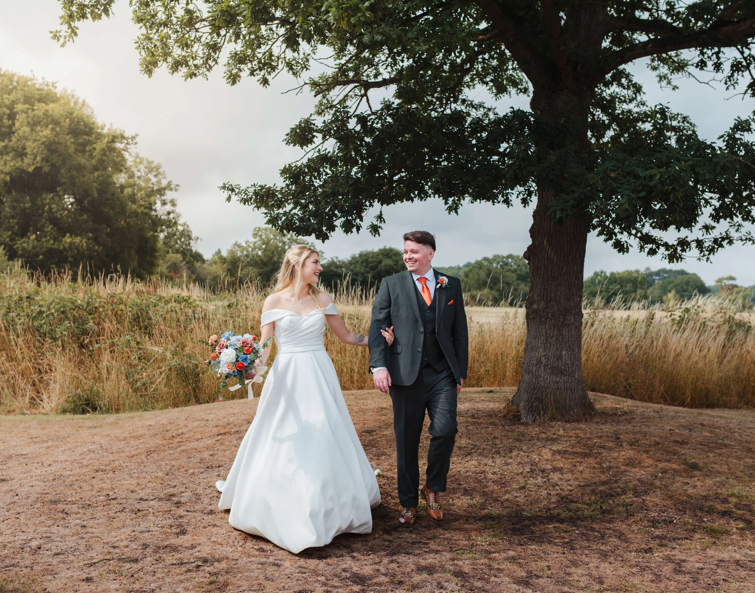 A bride and groom walk hand in hand outdoors under a large tree on their wedding day, smiling at each other.