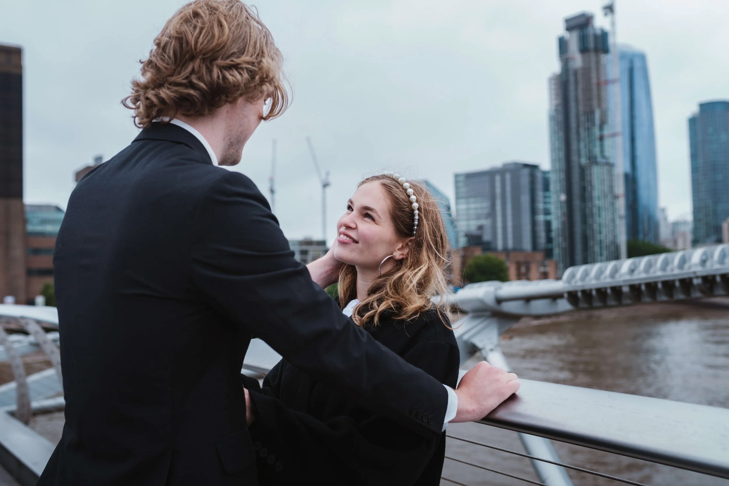 A young couple with light skin and curly hair standing close to each other on a city riverfront, with modern buildings in the background. The woman is smiling and looking at the man, who is gently holding her face.