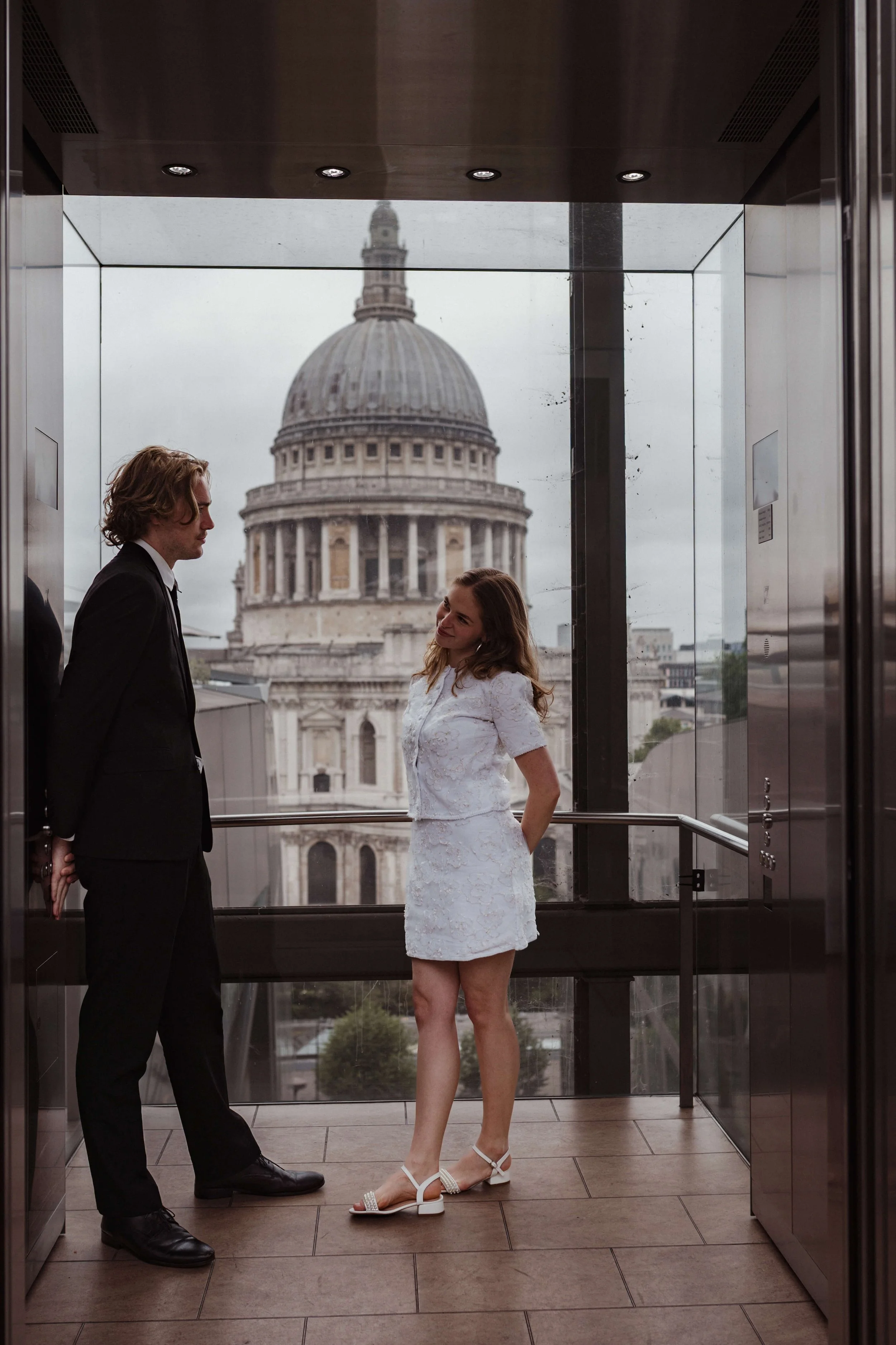 A man and a woman inside an elevator with a view of St. Paul's Cathedral in London through the glass window behind them. The woman is wearing a white dress and white sandals, smiling at the man. The man is dressed in a black suit, facing her.