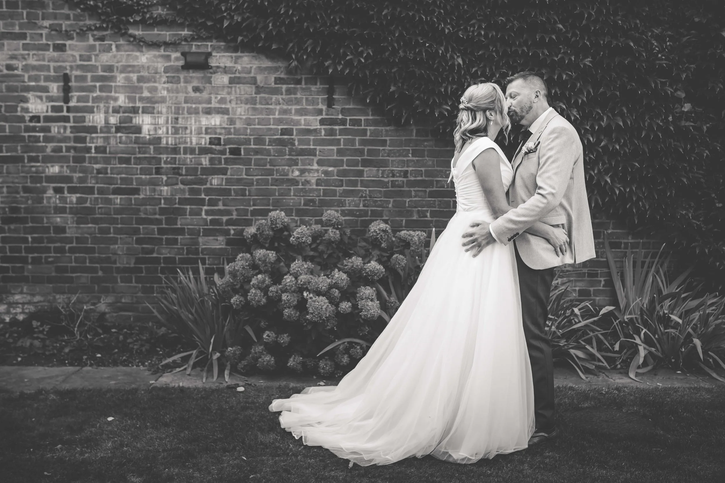 A black and white photo of a bride and groom standing closely together, about to kiss, outdoors in front of a brick wall with plants and flowers nearby.