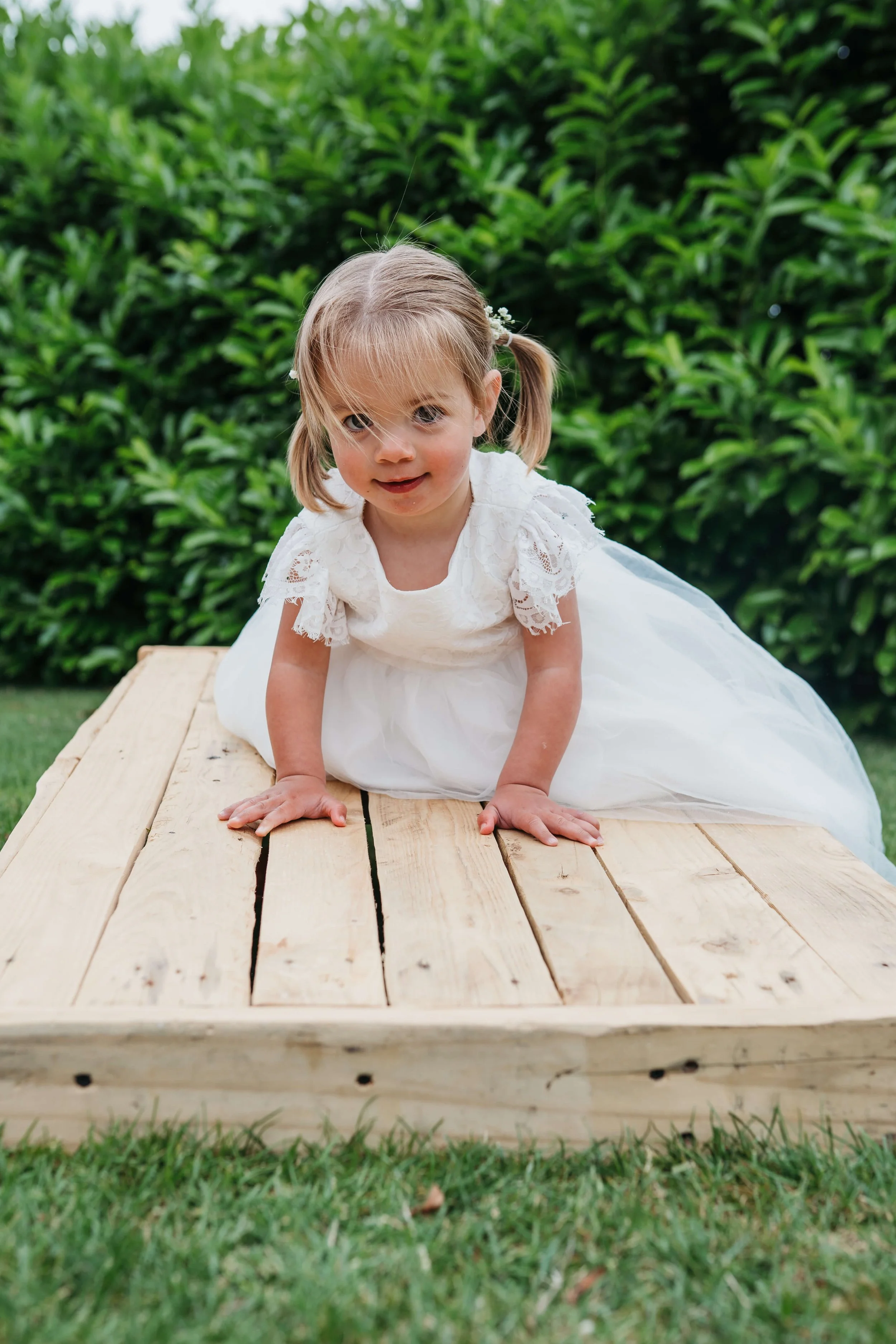 A young girl in a white dress crawling on a wooden platform outdoors with green bushes in the background.
