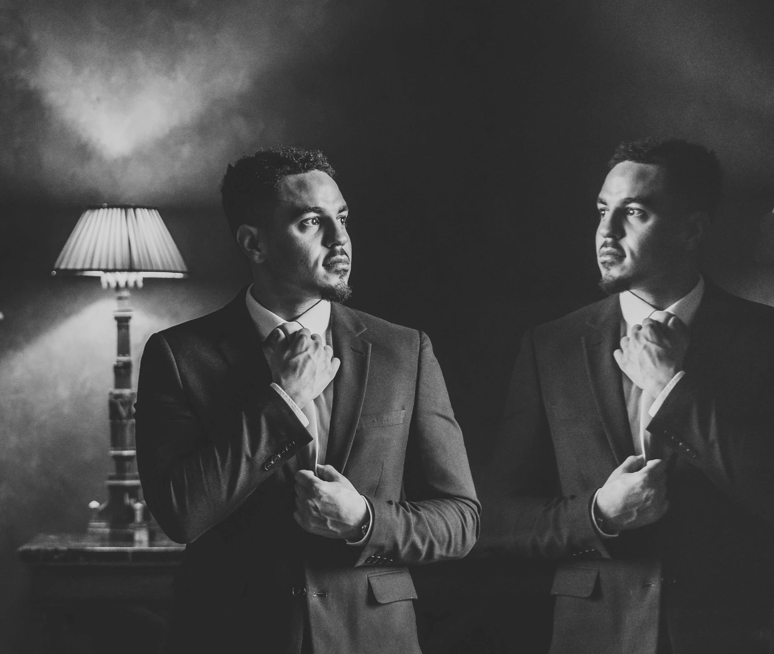 A black and white photo of a man looking at his reflection in a mirror, adjusting his tie, with a vintage table lamp in the background.