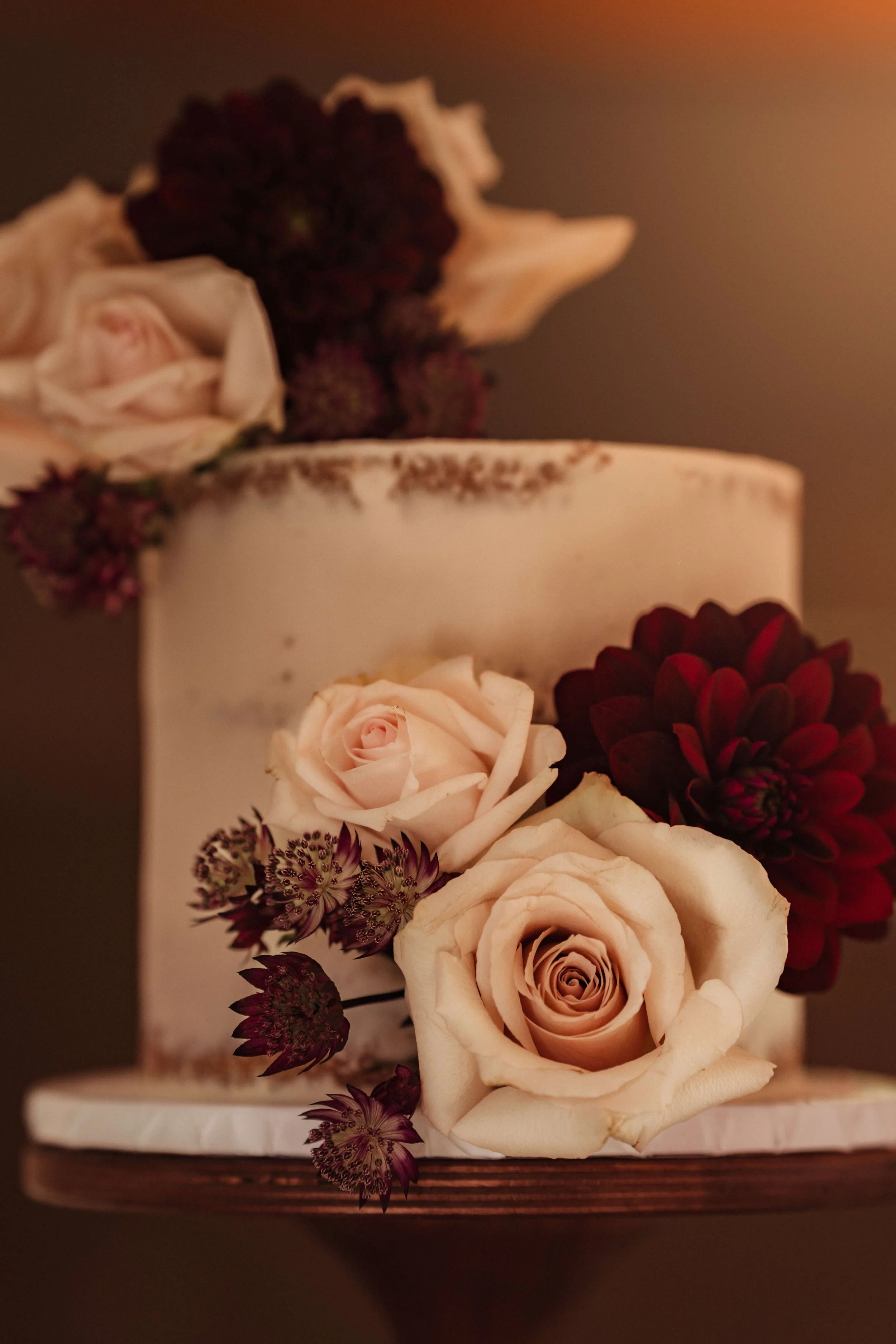 A cake decorated with blush pink roses and dark red dahlias, with sprigs of small dark purple flowers, on a wooden base.