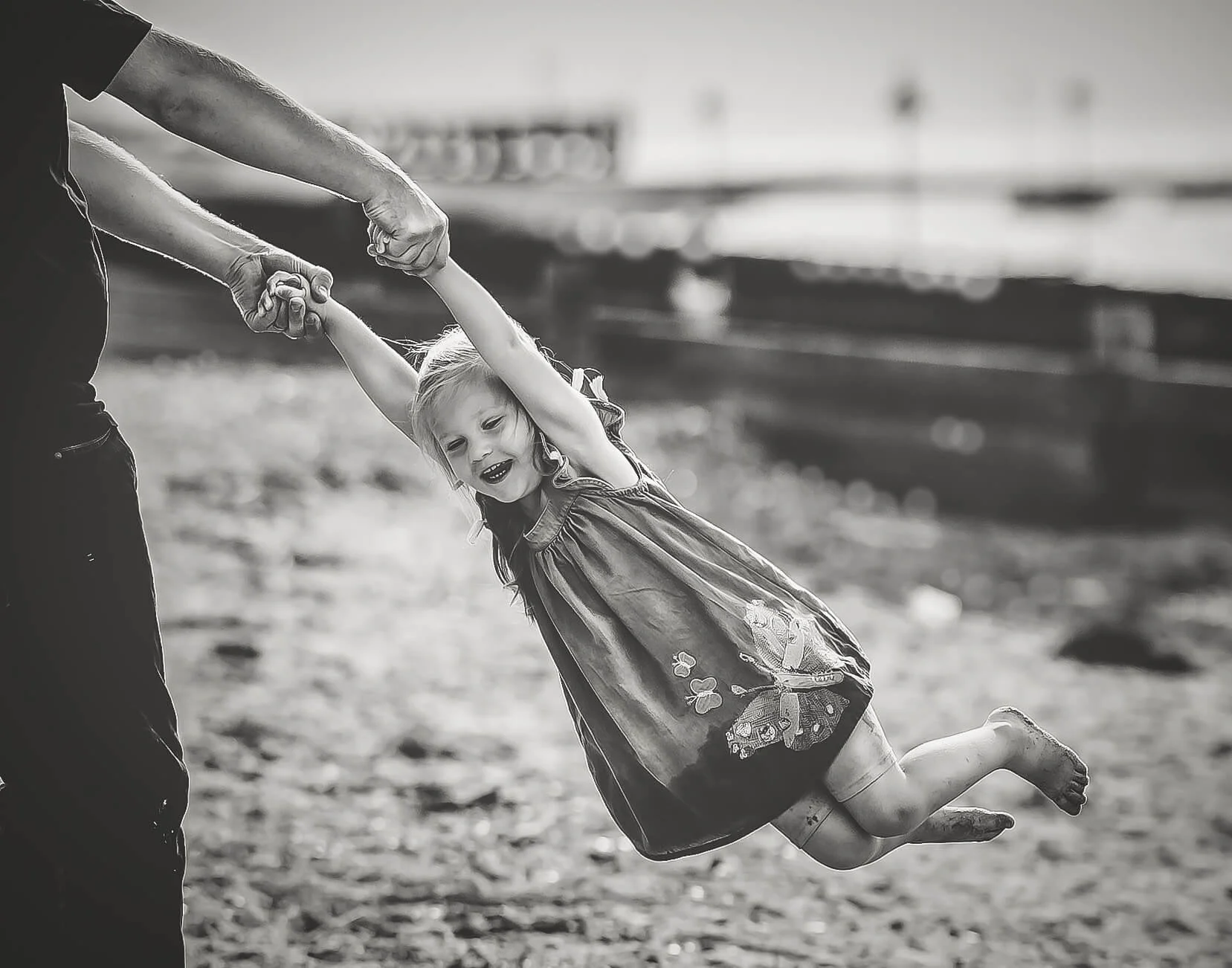 A young girl in a dress with butterfly embroidery is swinging while being held by an adult outdoors near a body of water.