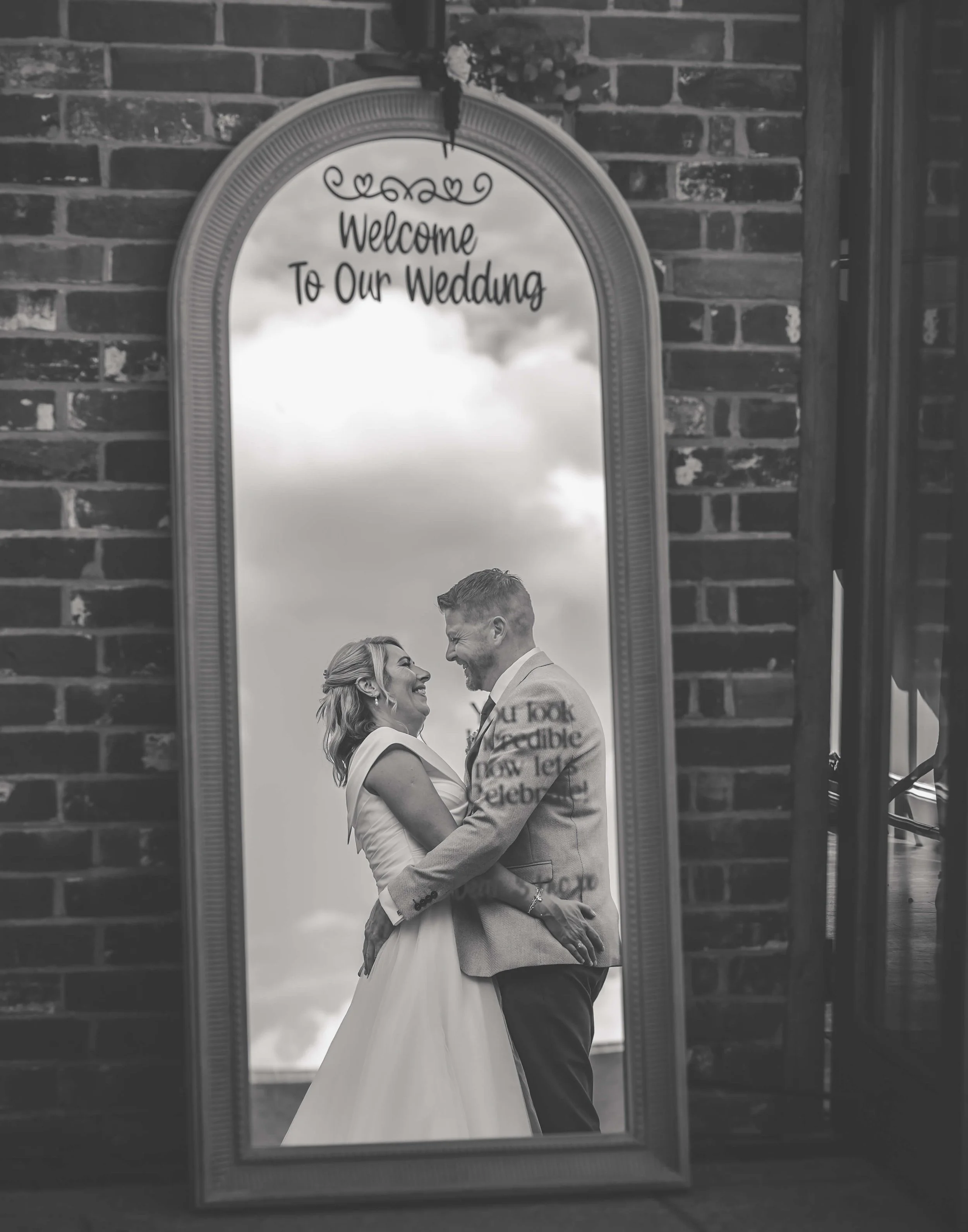 A black-and-white photo of a bride and groom looking at each other and smiling, seen through a tall, arched mirror with a message at the top that says "Welcome to Our Wedding". The background features clouds and the setting appears to be indoors with