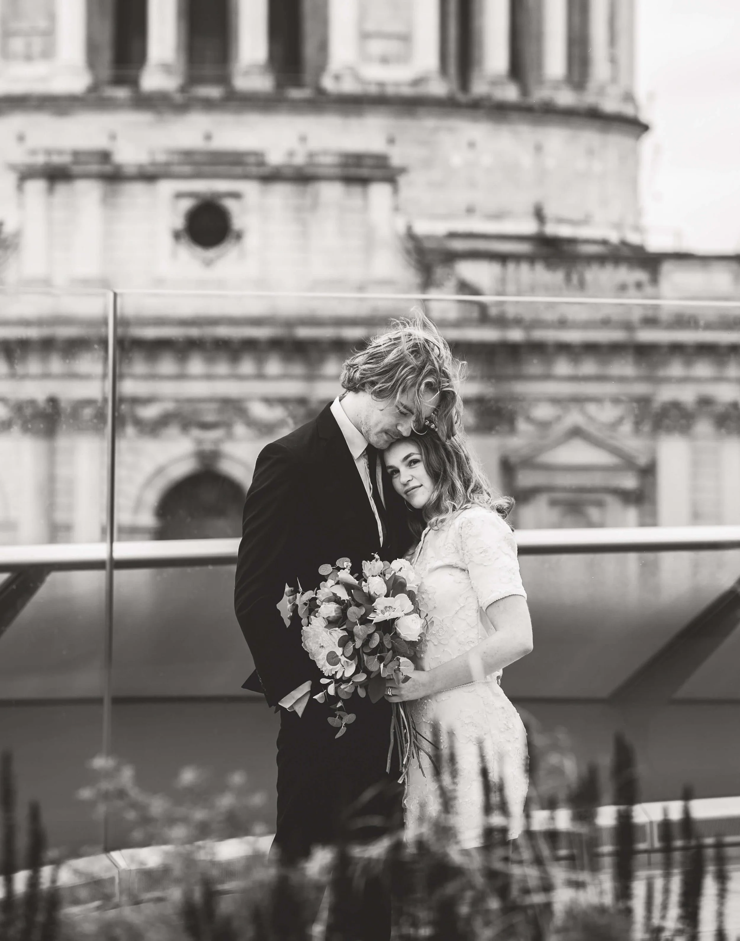 A black and white photo of a bride and groom embracing, with the bride holding a bouquet of flowers, standing on a balcony with a historic building in the background.