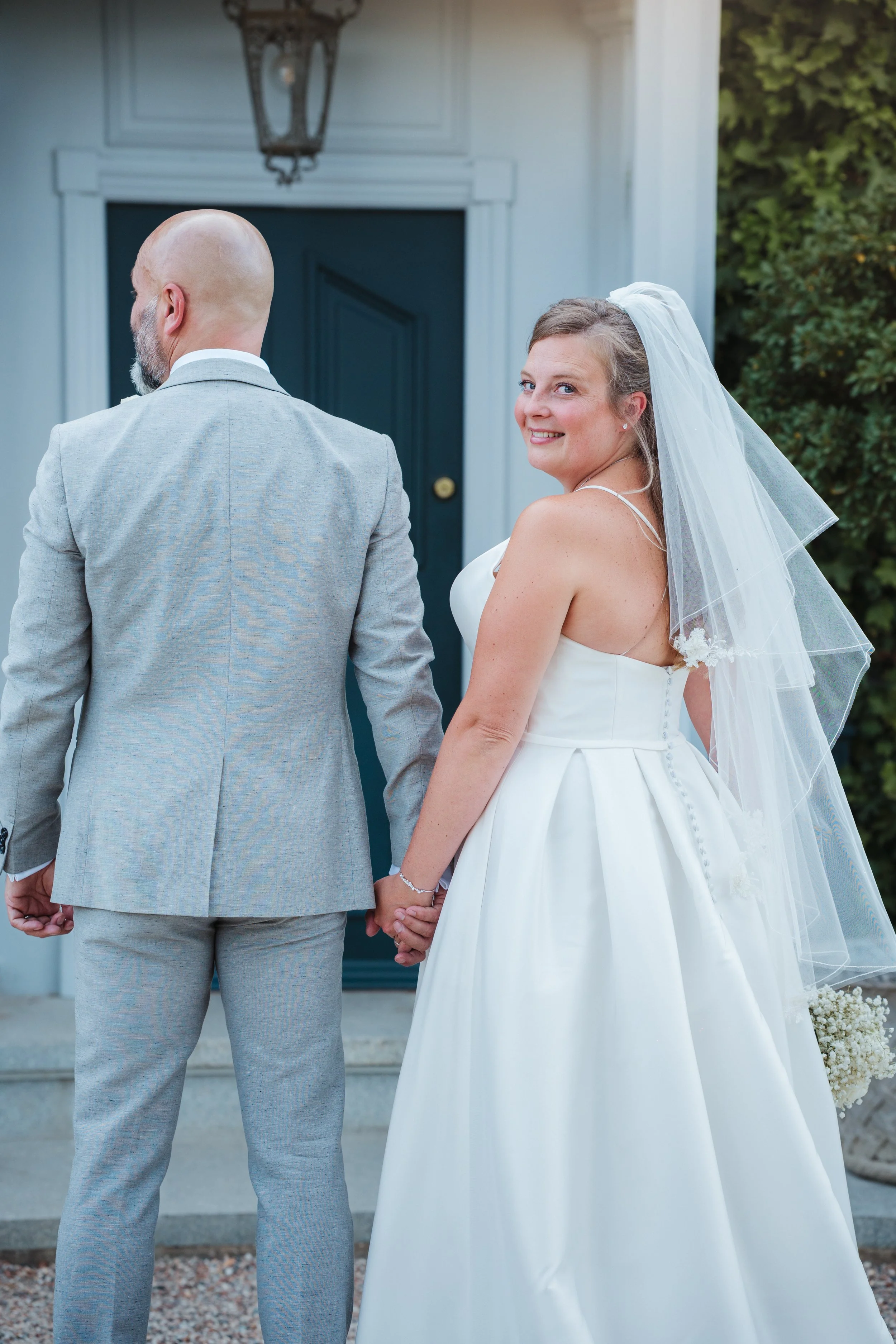 A bride in a white wedding dress and veil holding hands with her groom in a gray suit, smiling and looking at the camera outside a house.