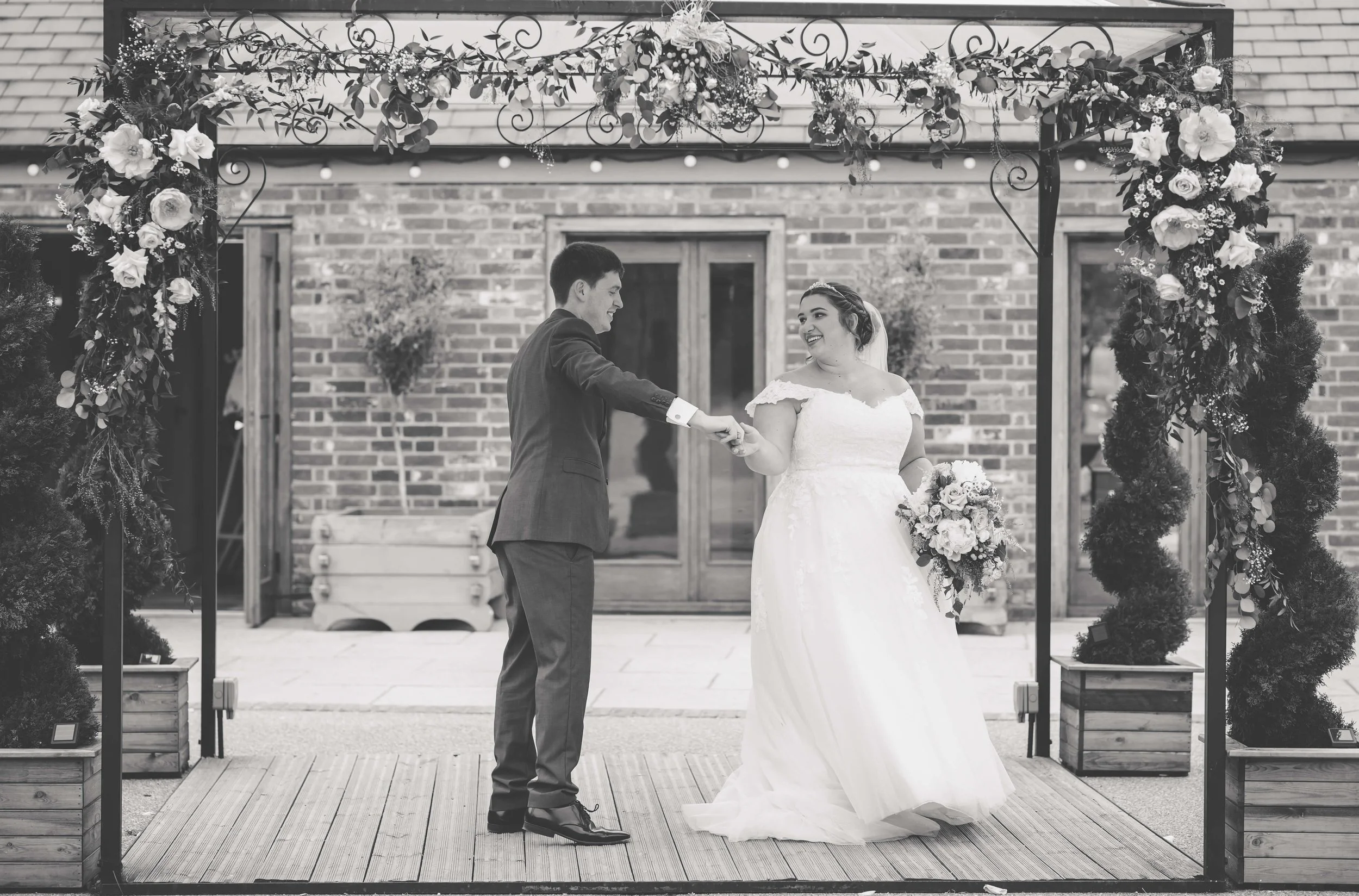 A bride and groom holding hands and smiling at each other under a decorated archway during their wedding ceremony outside, with a brick building in the background.